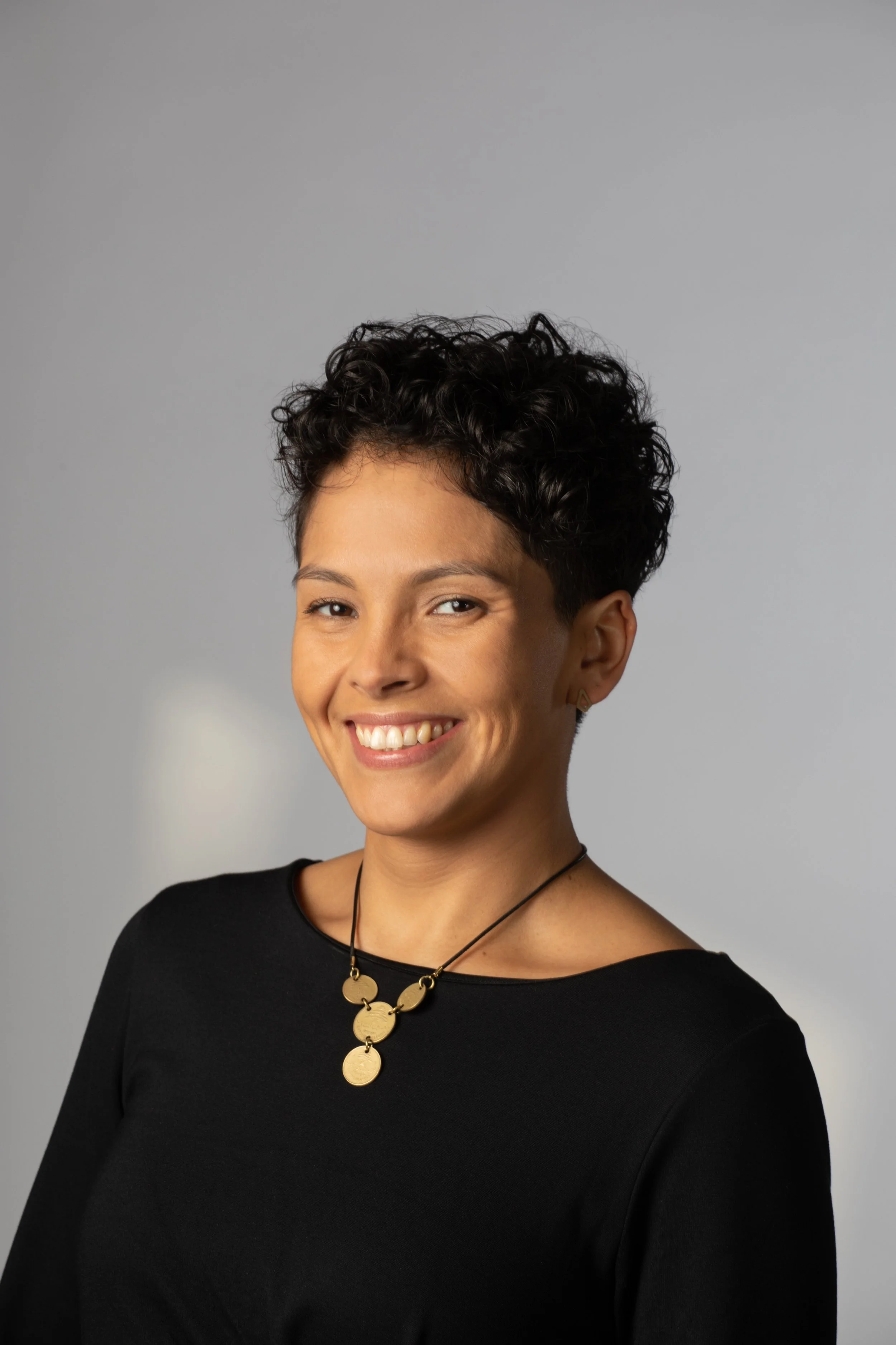 Portrait of a smiling woman with short curly dark hair wearing a black top and gold jewelry against a light gray background.
