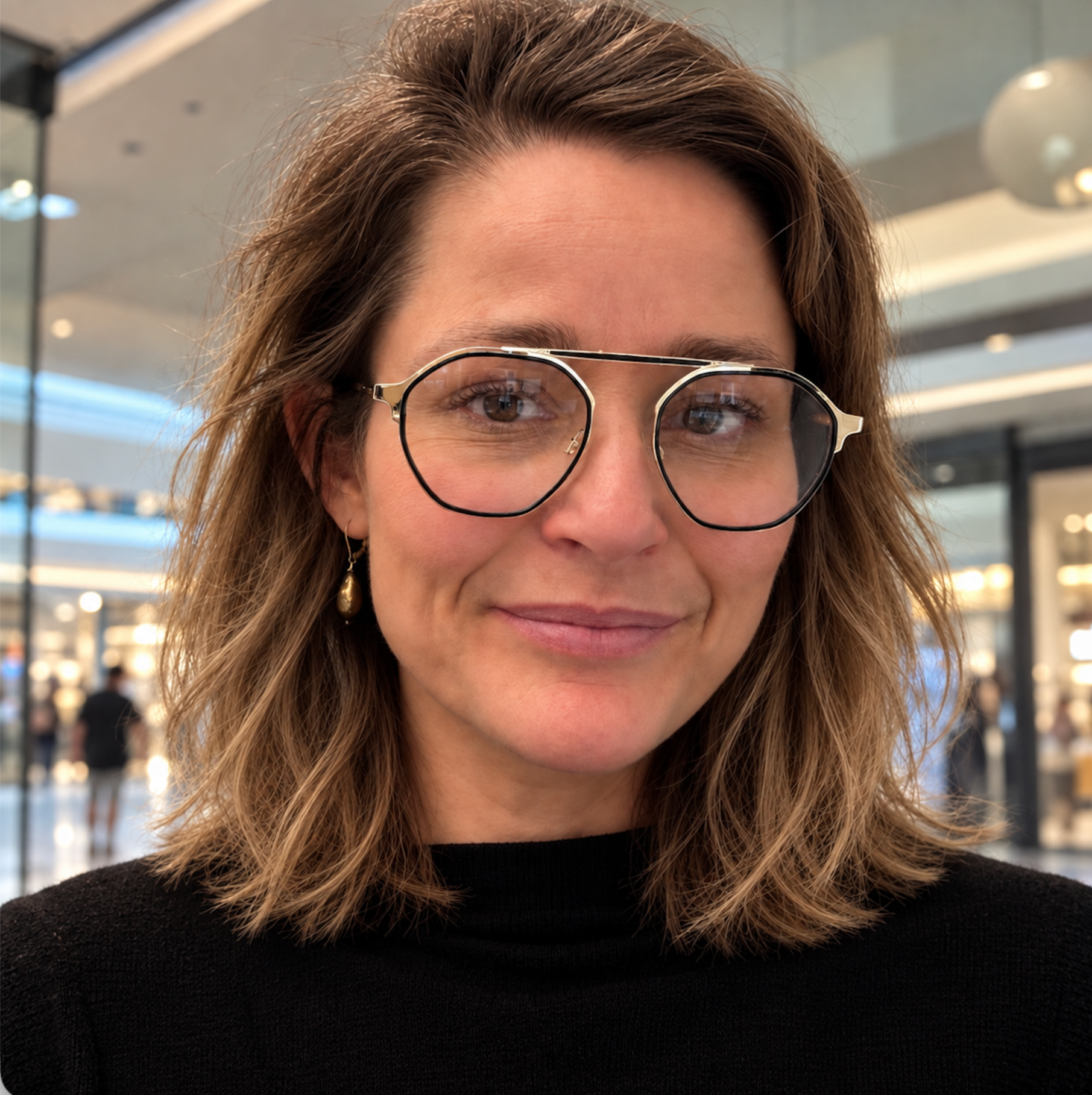 Close-up of a woman with brown, wavy hair, glasses, and a black top, smiling slightly, in an indoor setting with blurred background.