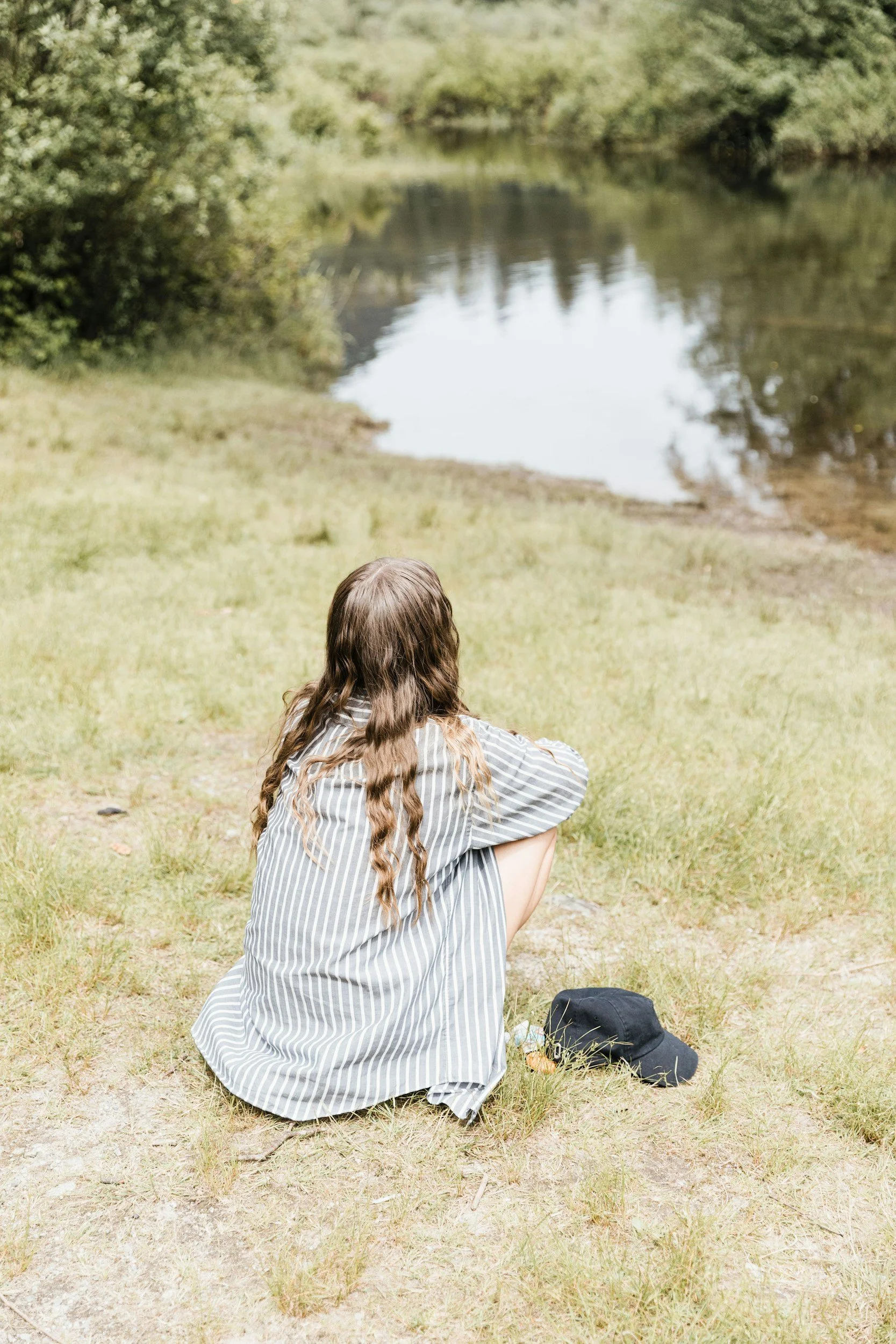 A person with long brown hair, wearing a striped shirt, sitting on the grass by a river, facing away from the camera, with a black cap on the ground nearby.