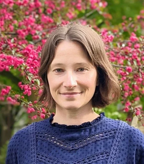 A woman with short brown hair wearing a blue embroidered blouse, standing outdoors in front of pink flowering bushes.