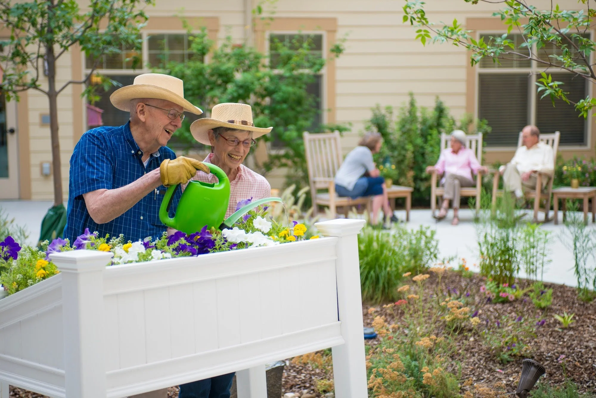Older man and woman smiling while watering flowers in a garden planter, with three other people sitting on chairs in the background in a backyard garden.