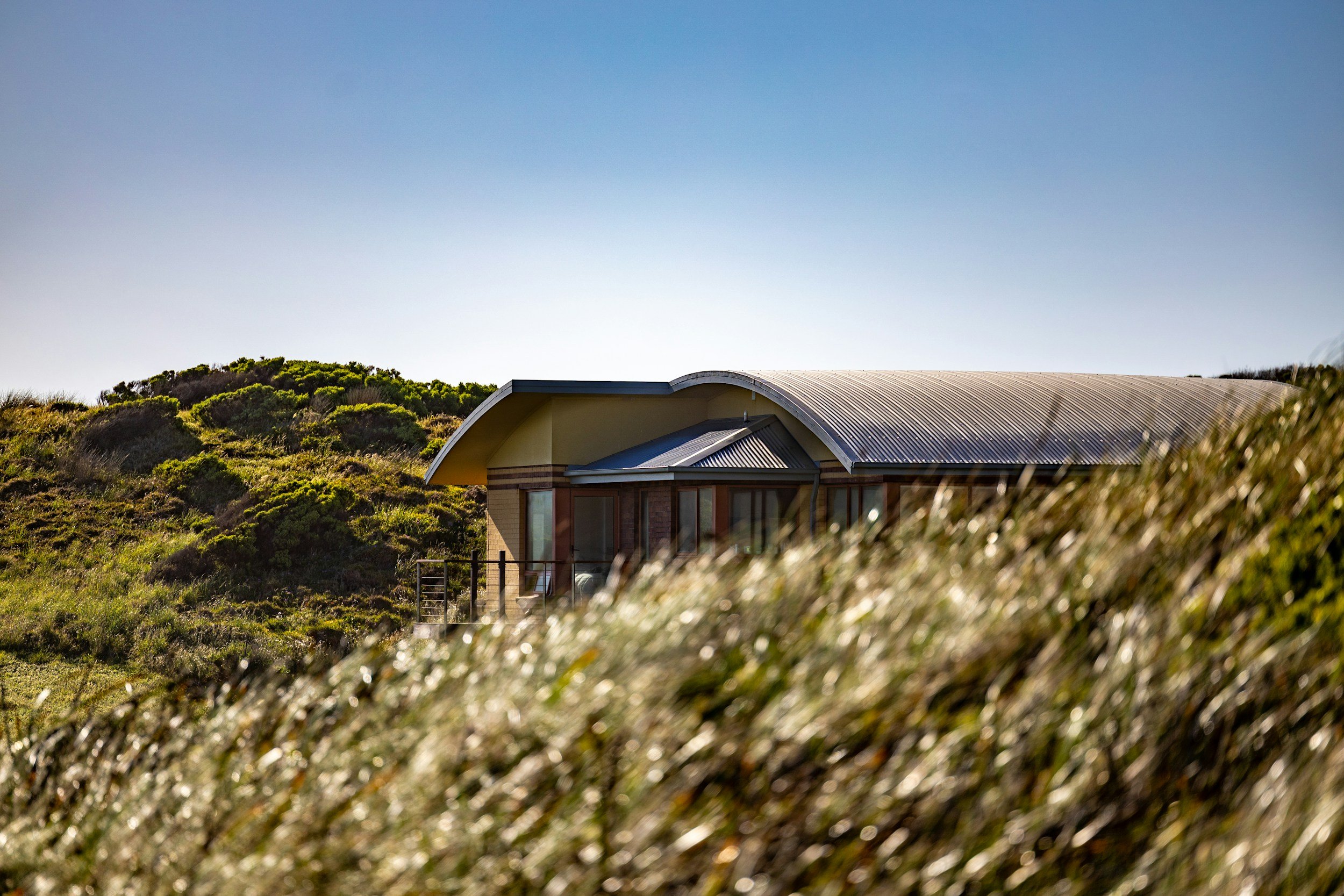 An elevated single story home with convex roof lines, earth tones exterior plaster, nestled into the hills of grasslands