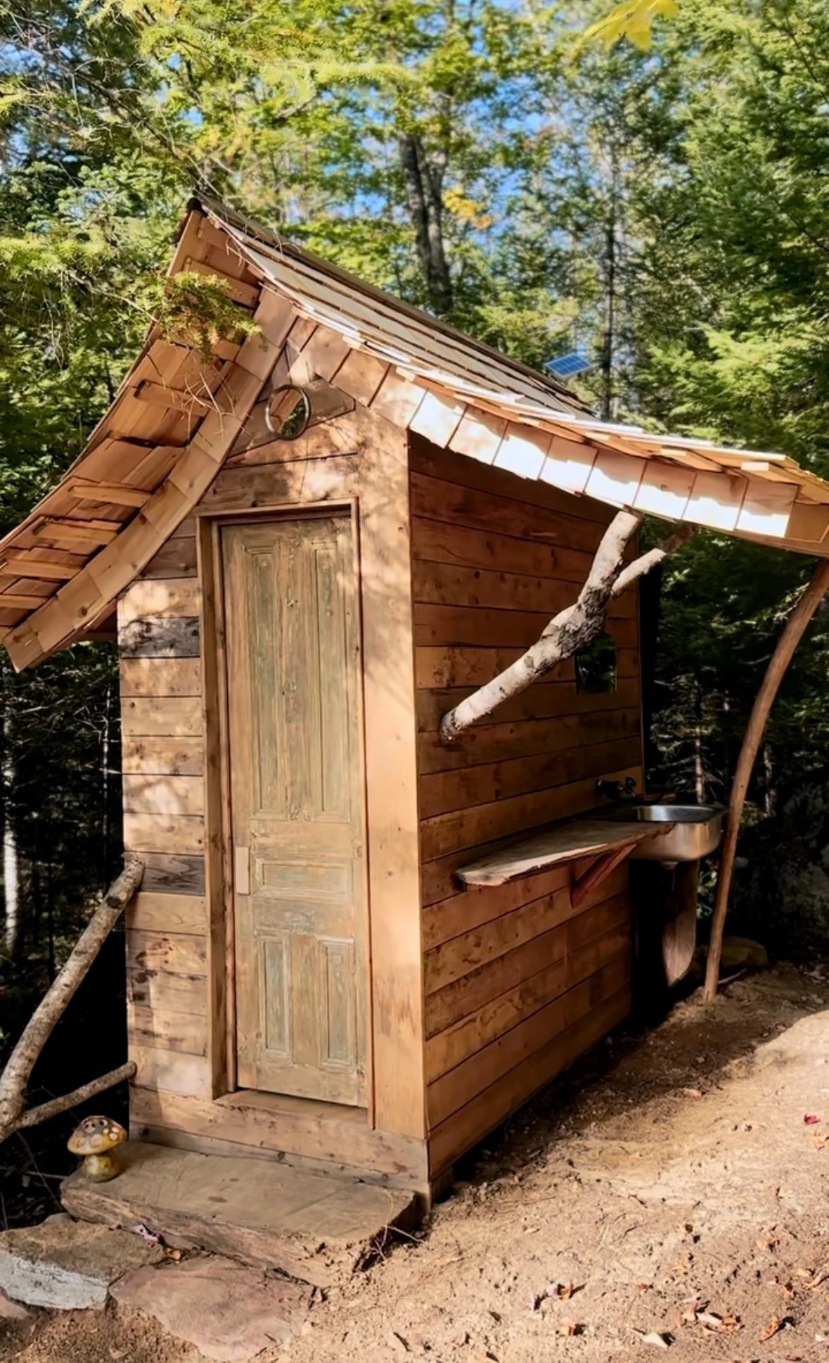 Toilette extérieure artisanale d’inspiration féerique et witchy, avec toit en bardeaux de cèdre et branches comme supports.