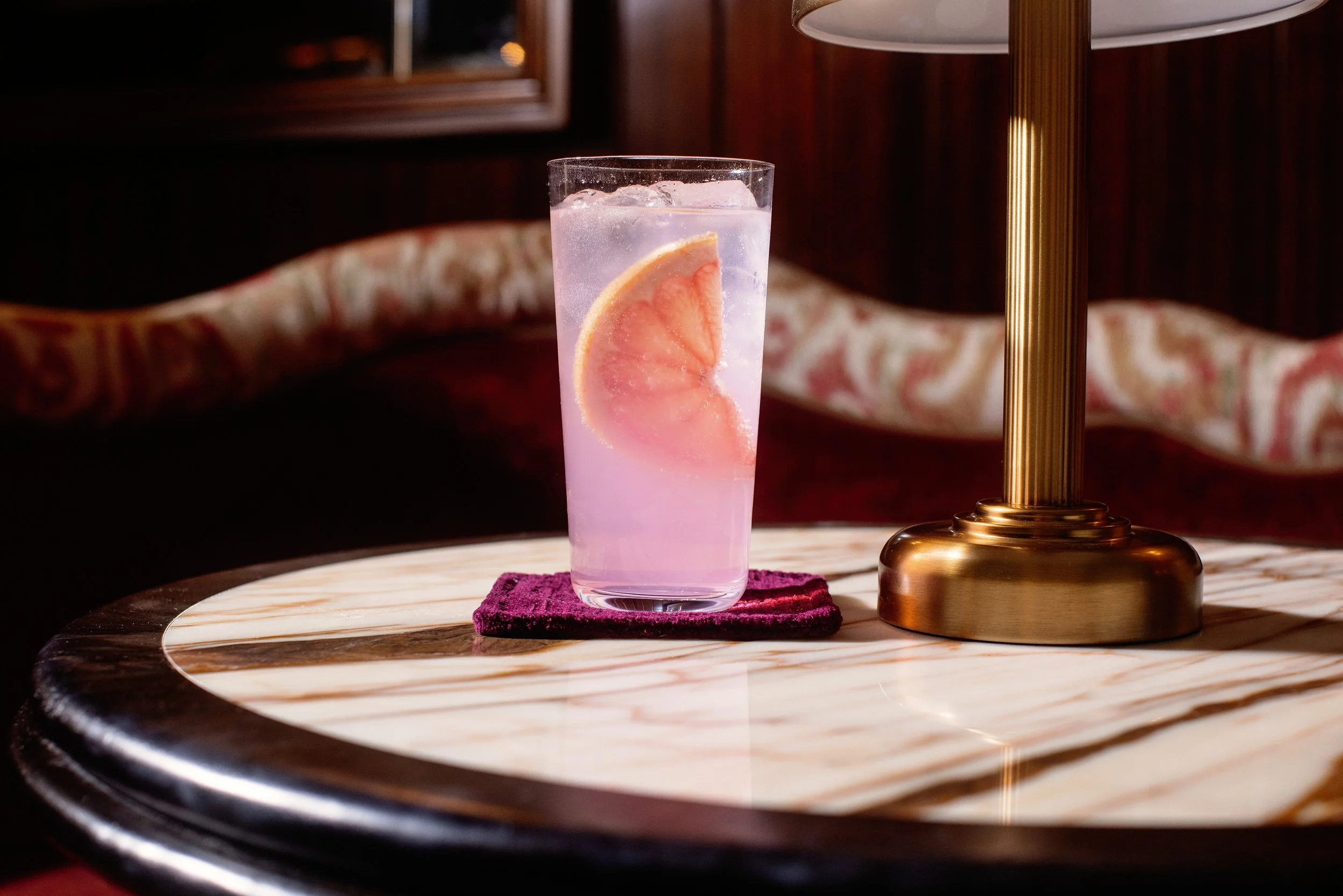 A tall glass filled with a pink lemon drink and ice, garnished with a lemon wedge, sitting on a purple cloth on a marble table, next to a gold-colored lamp in a dimly lit room with a patterned sofa in the background.