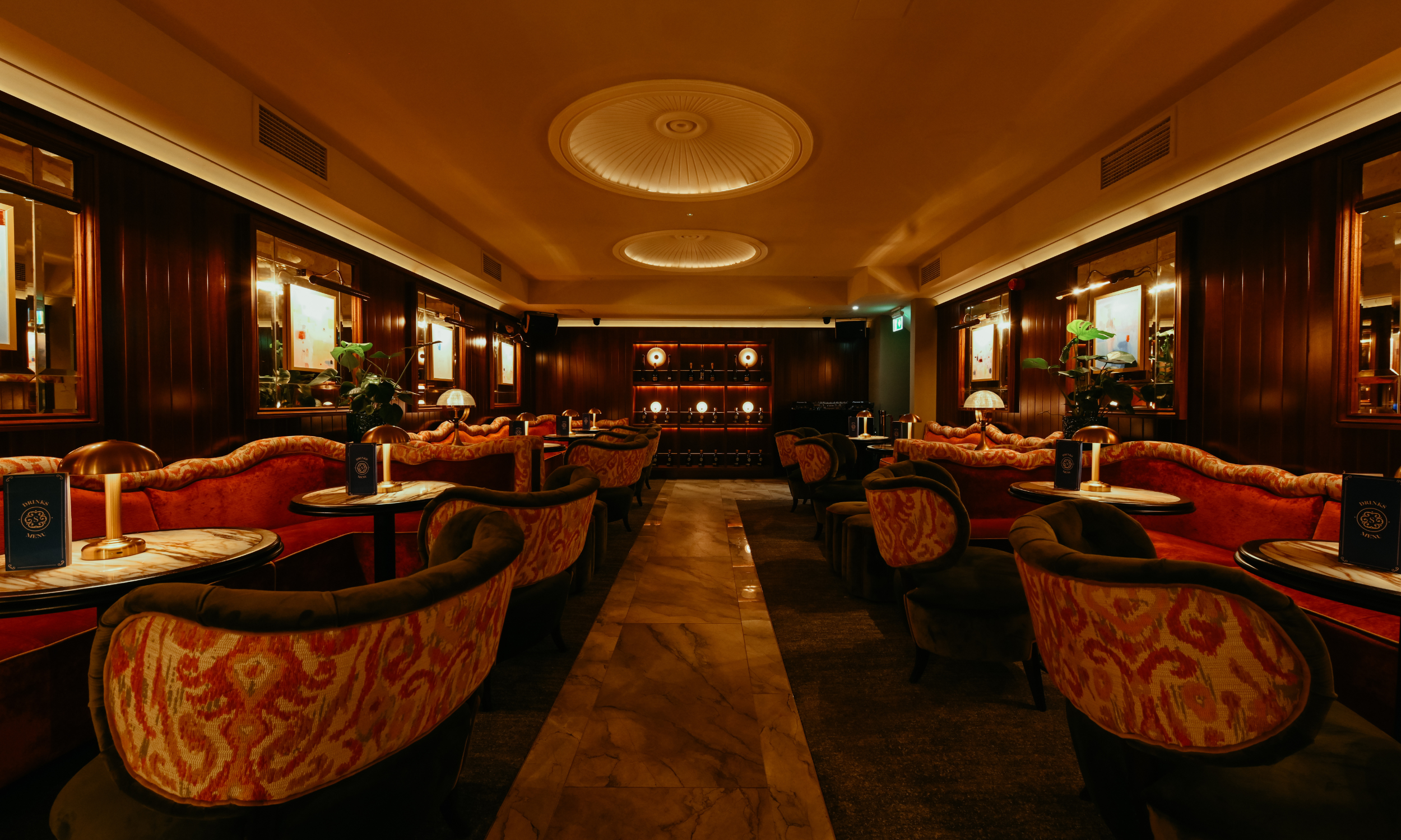 A luxurious, dimly lit lounge with plush, patterned red and gold chairs and sofas arranged around marble-topped tables. Dark wooden wall panels and framed artworks line the walls. Small table lamps provide warm lighting. The back wall features decora