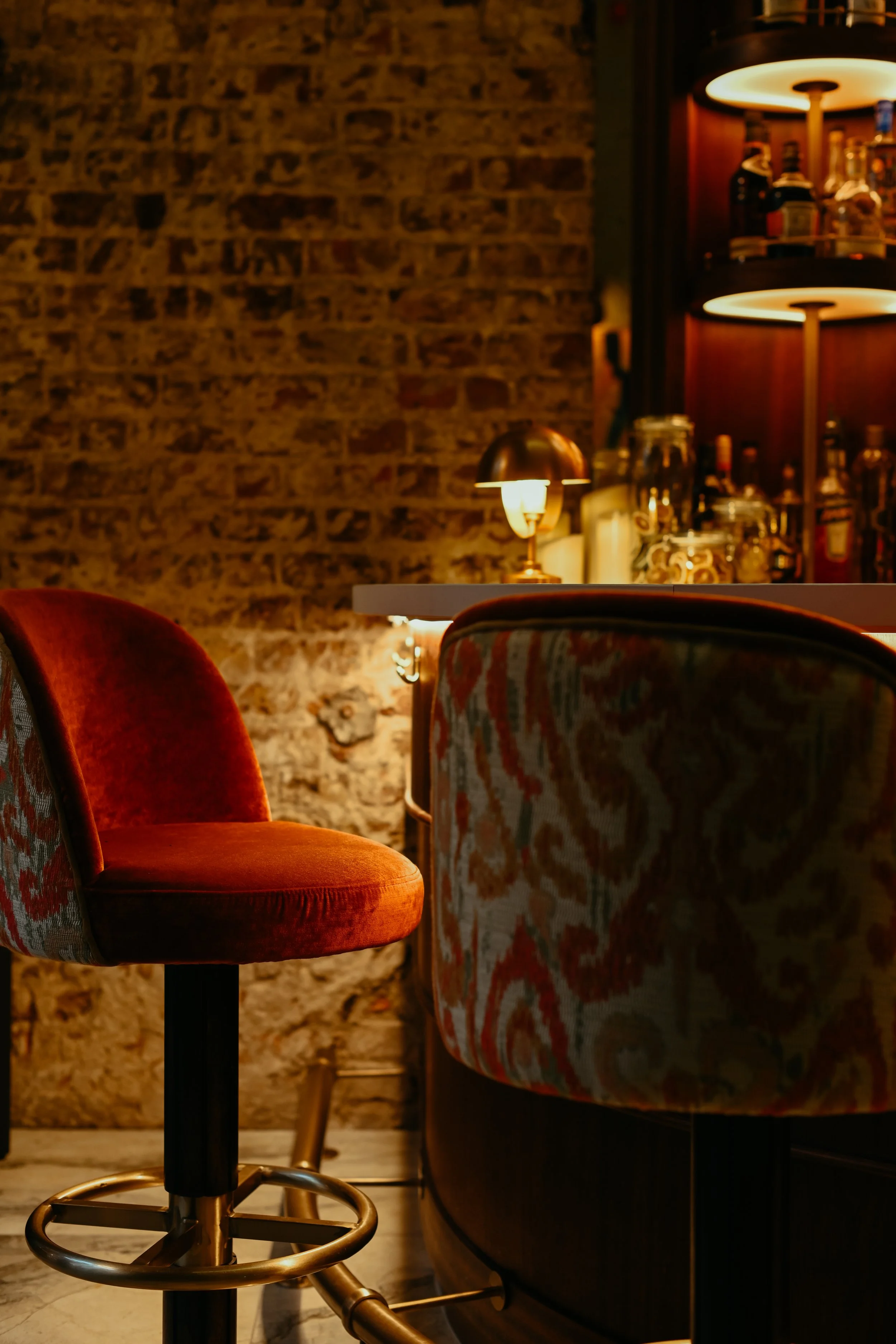 A cozy bar interior with a brick wall, a plush red velvet bar stool, and a patterned upholstered chair near a white counter. Warm lighting from a small lamp and wall sconces creates an inviting atmosphere.