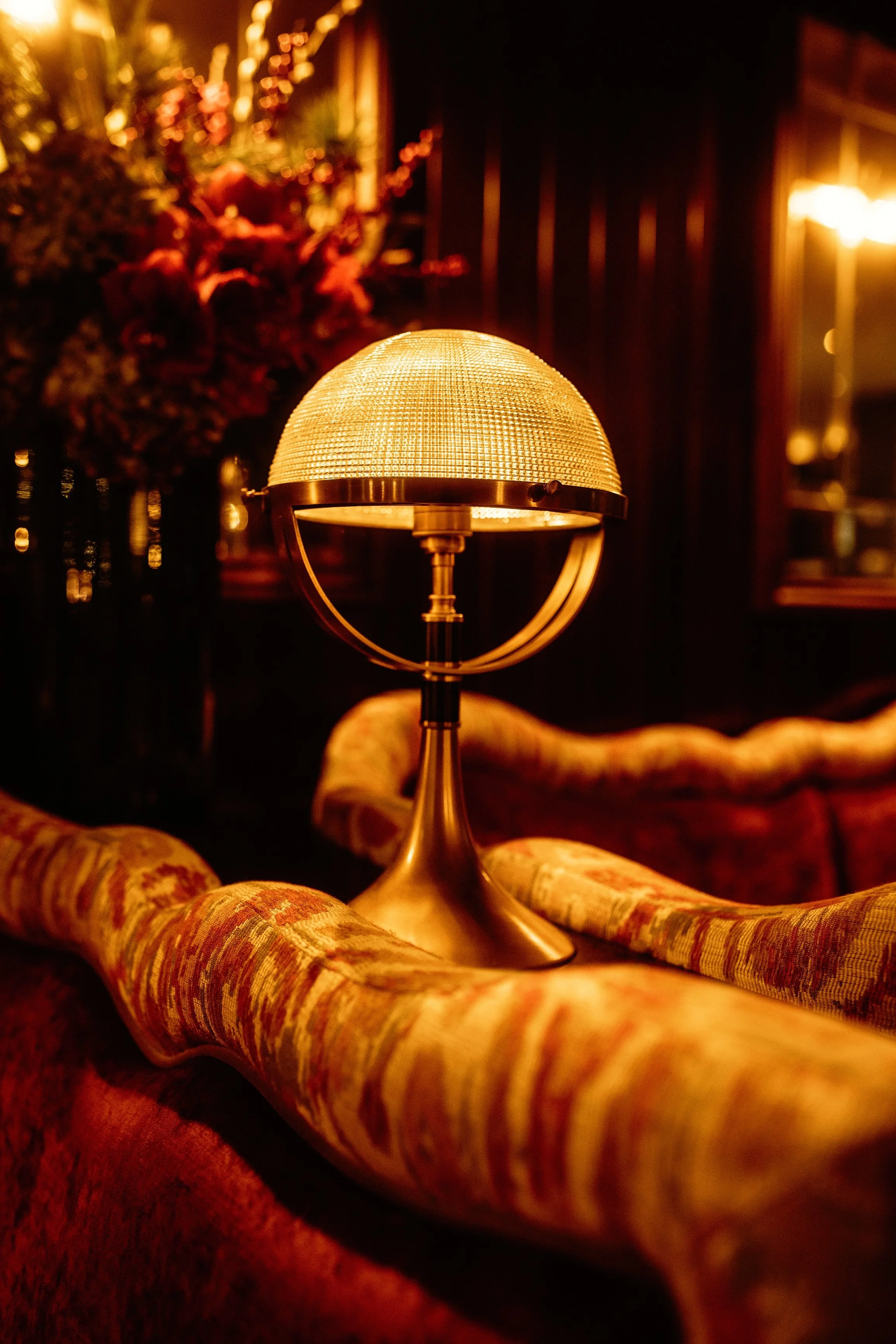 A vintage-style table lamp with a dome-shaped shade sitting on a patterned fabric surface in a dimly lit room, with a blurred floral arrangement and warm lighting in the background.