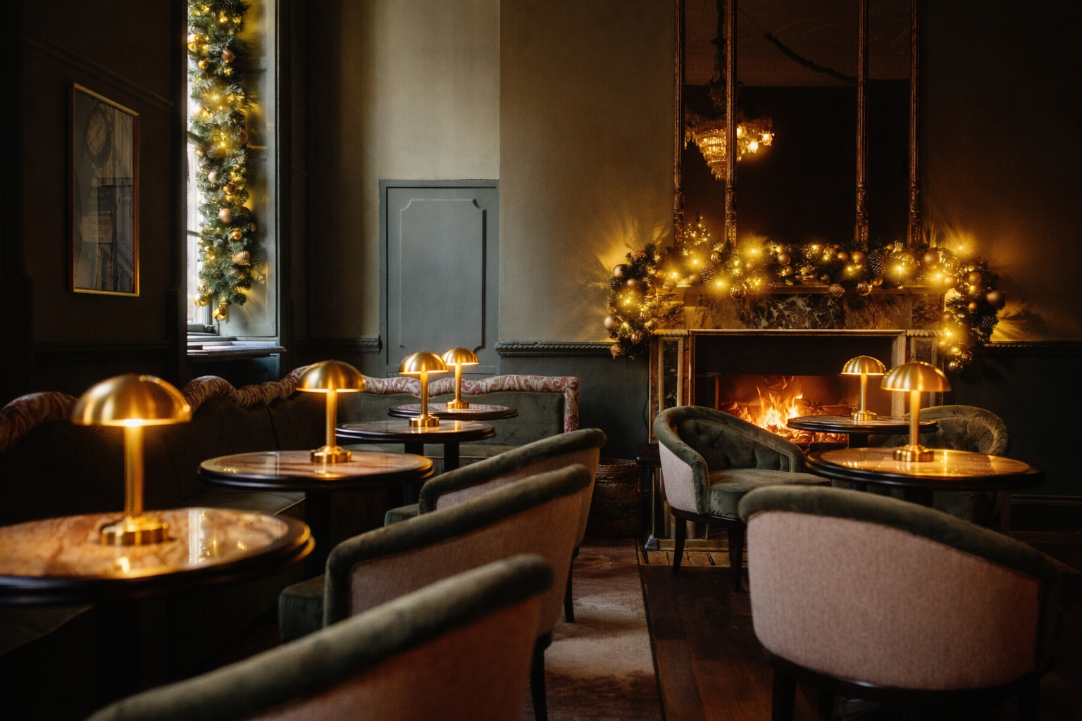 Cozy, dimly lit room decorated for Christmas with a fireplace, gold garland, and Christmas ornaments. Small tables with gold lamps and upholstered chairs are arranged around the room.