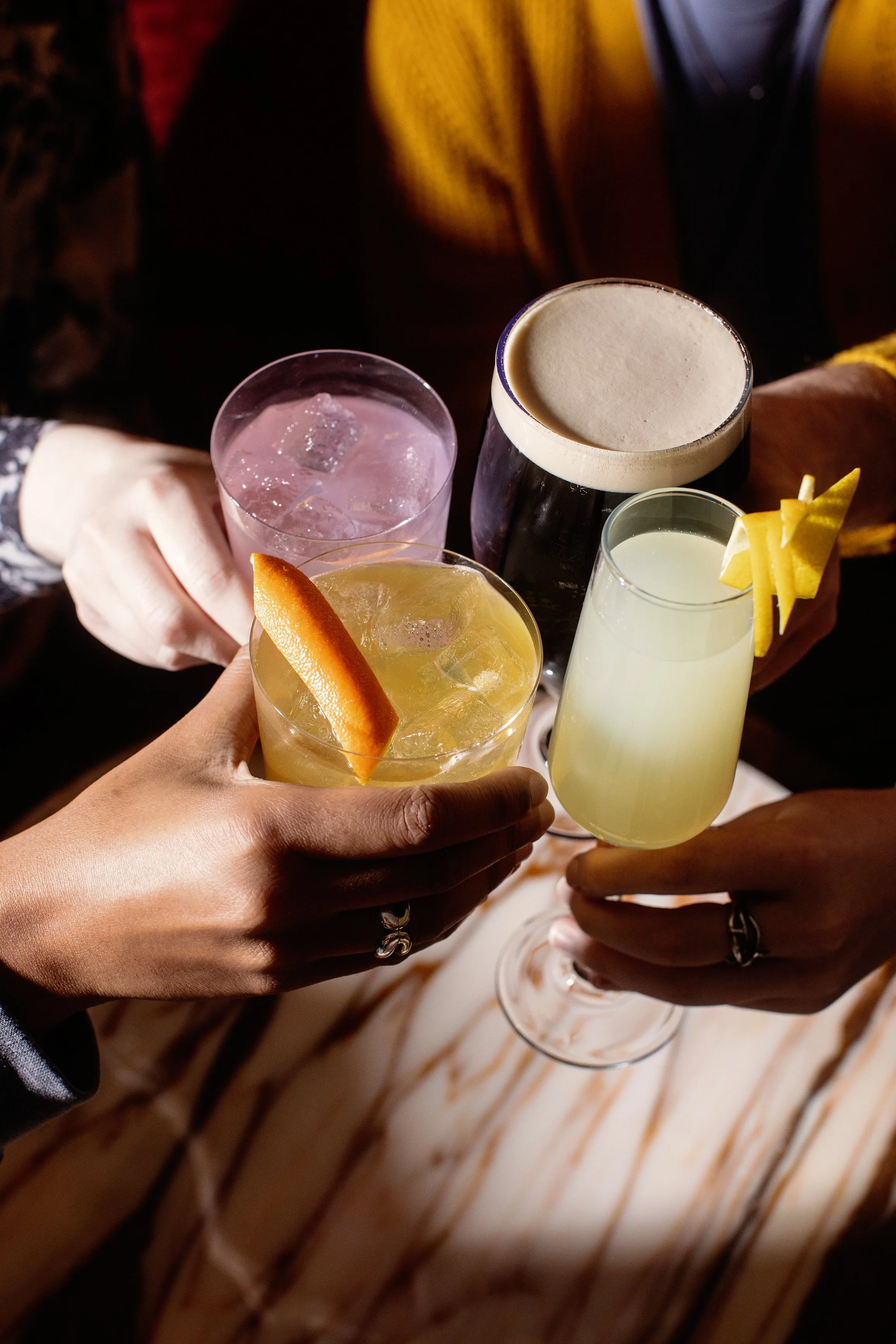 Group of people holding various cocktails in glasses for a toast, including a dark beer, a pink cocktail, a yellow cocktail with an orange slice, and a light-colored bubbly drink with lemon peel.