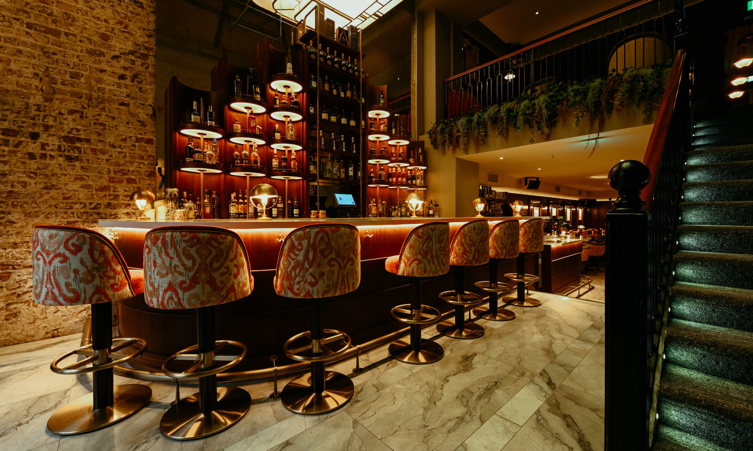 Elegant bar with a curved marble countertop, velvet upholstered bar stools, illuminated liquor shelves, and ambient lighting, with a staircase on the right and a brick wall on the left.