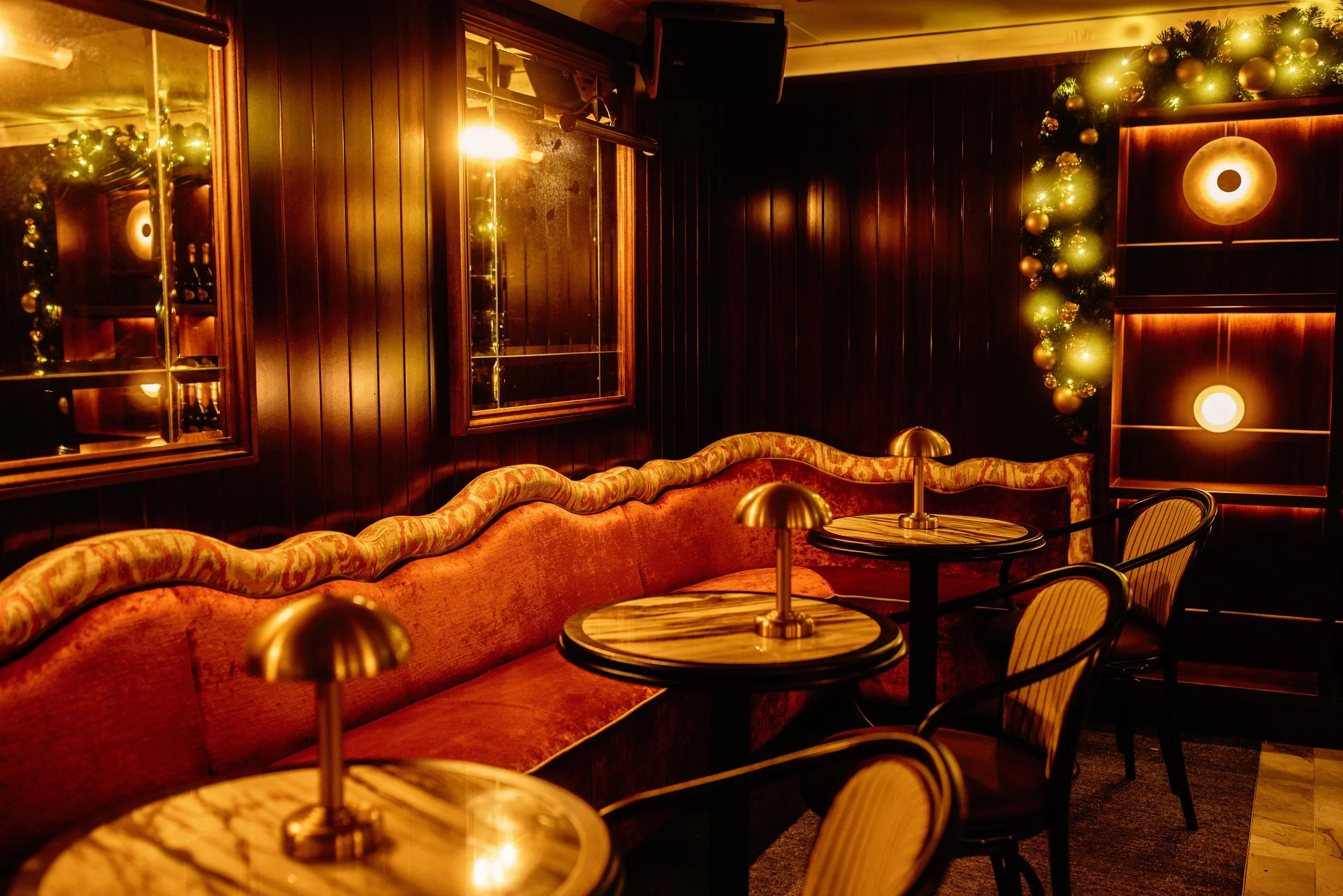 A cozy, dimly lit bar or lounge decorated for the holidays with a Christmas wreath and Christmas ornaments on the wall, velvet seating, small tables with brass lamps, and dark wood paneling.