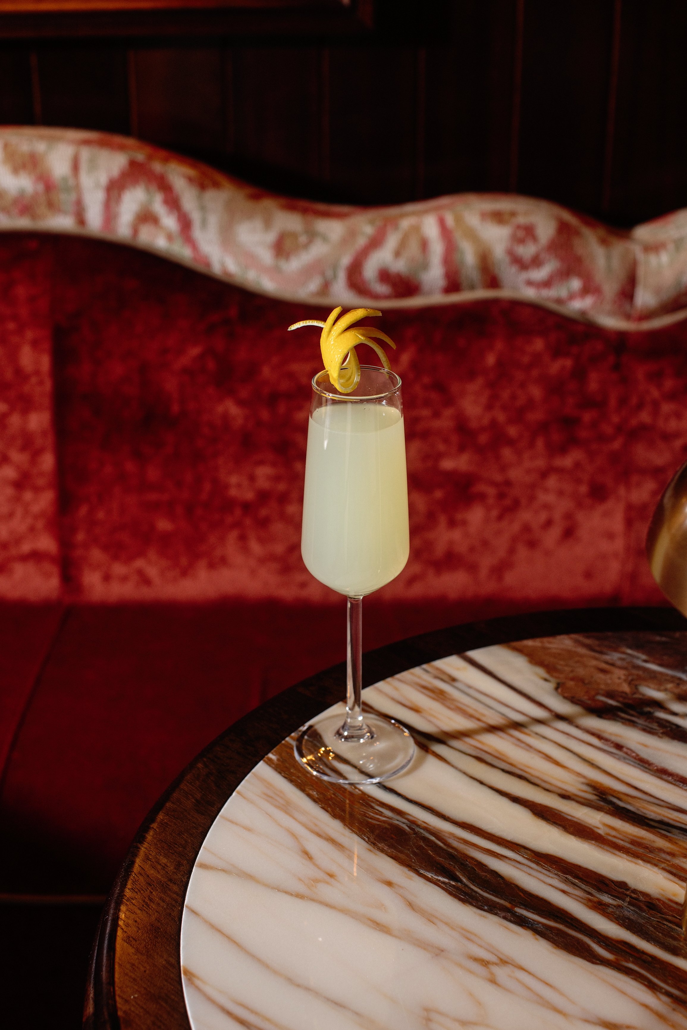 A champagne glass filled with a light-colored cocktail, garnished with a lemon peel twist, sits on a marble-topped table in a restaurant or lounge setting.