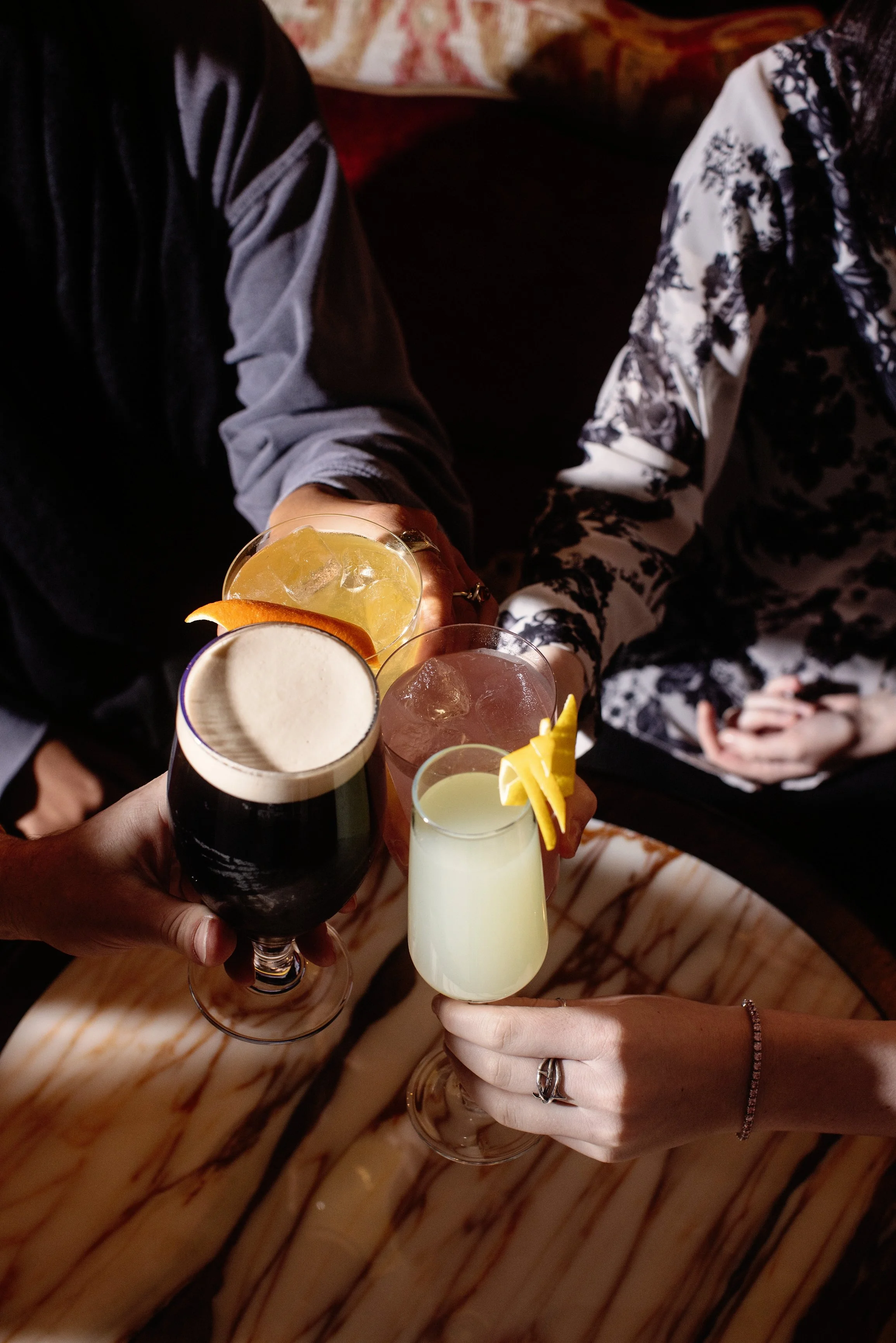 People toasting with various cocktails at a bar, showing hands and drinks with garnishes on a wooden table.