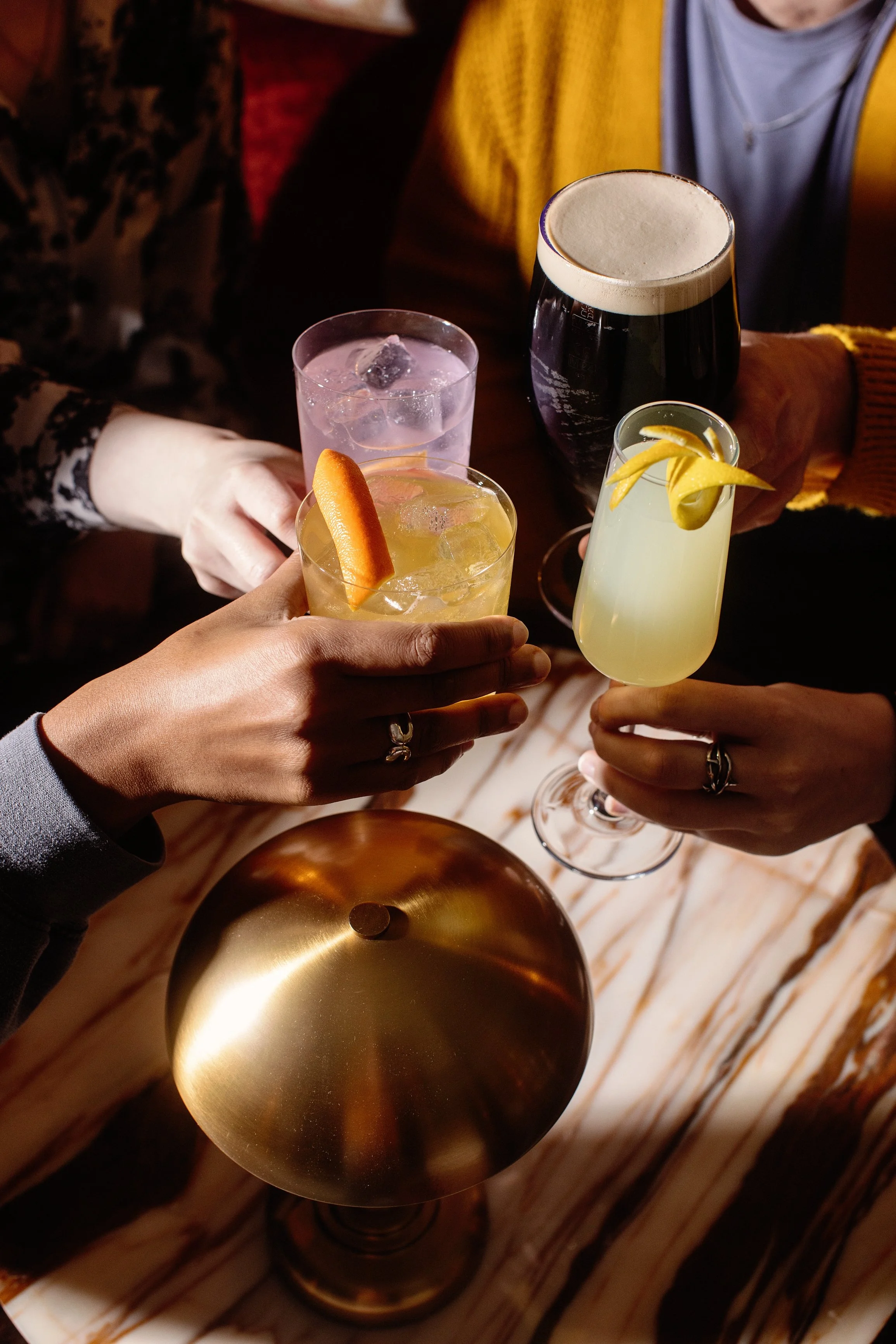 A group of people holding various cocktails in a dimly lit setting, with a golden candle holder on a marble table.