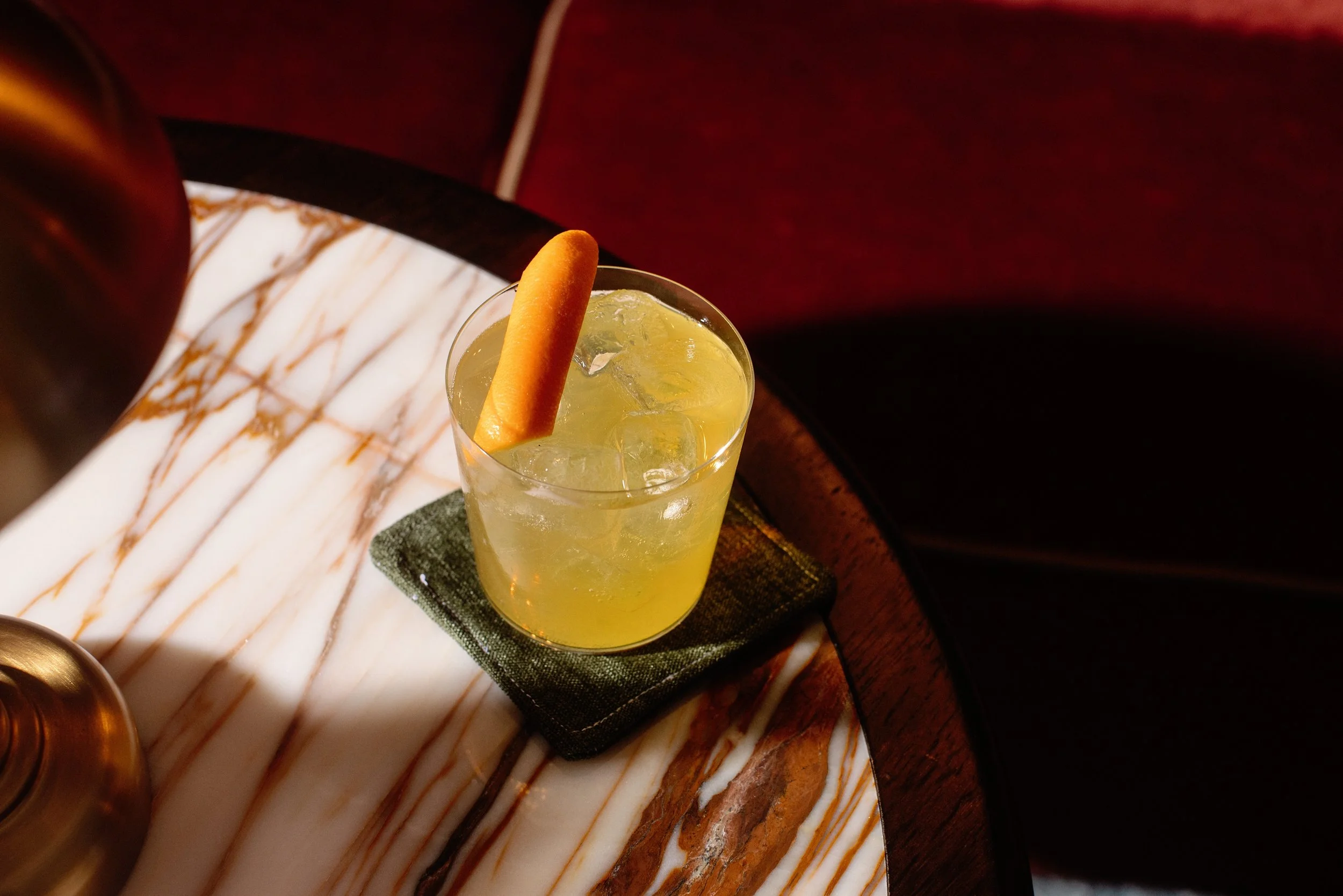 A yellow cocktail with ice and a lemon wedge in a glass, placed on a black coaster on a marble table with a red background.