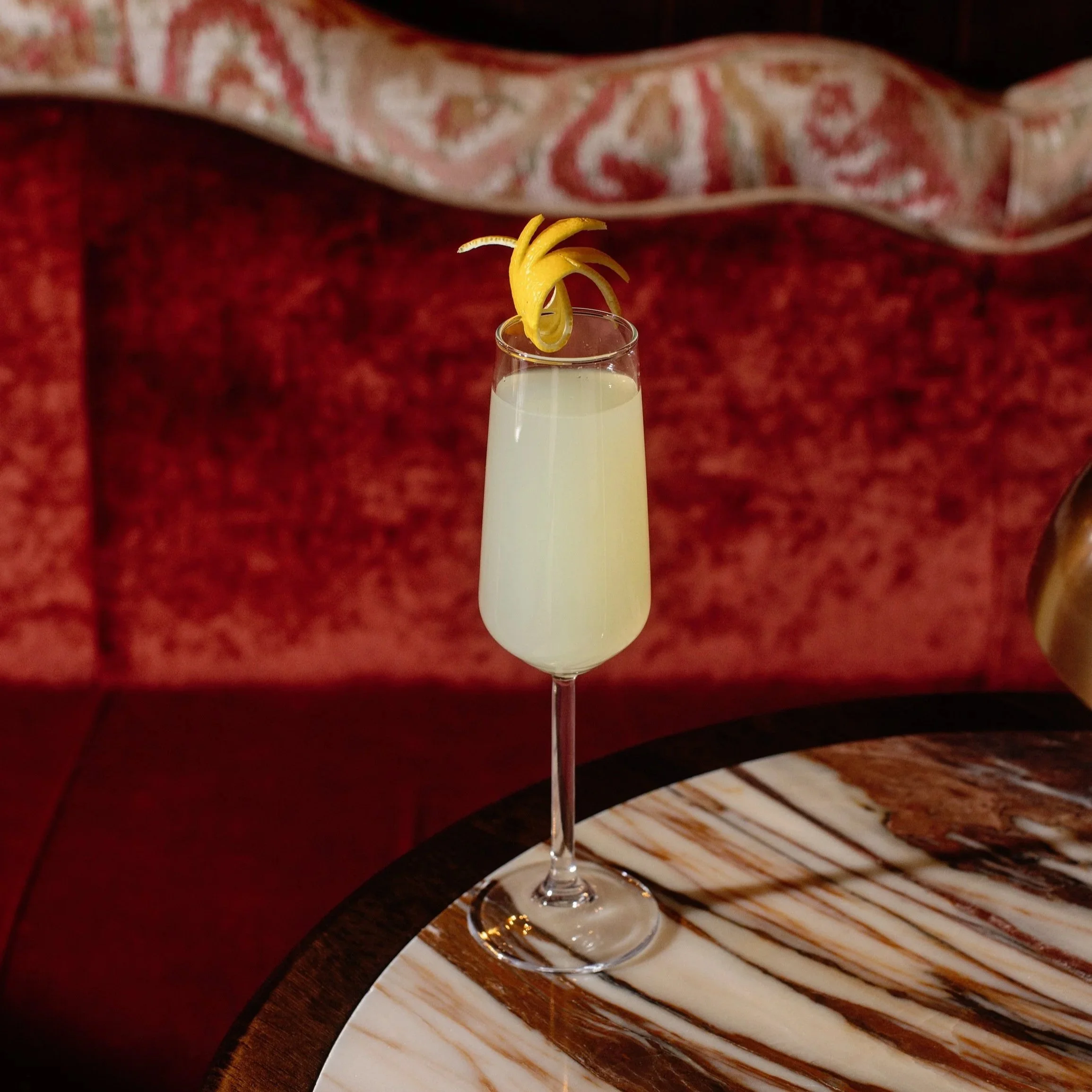 A champagne flute filled with a light-colored cocktail, garnished with lemon peel twists, on a marble-topped wooden table in a cozy, dimly lit setting.