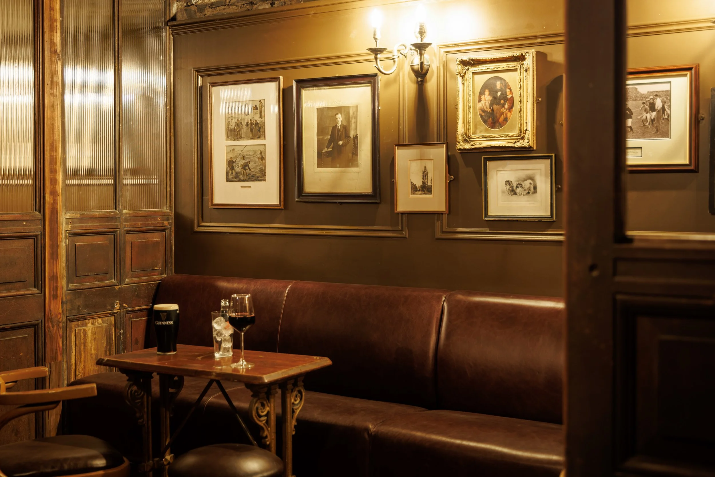Interior of a cozy pub or bar with a dark wooden wall decorated with framed black-and-white and color artwork, a leather banquette seating, a small wooden table with a glass of red wine, a pint of Guinness, and a glass of ice water, illuminated by a wall-mounted light fixture.