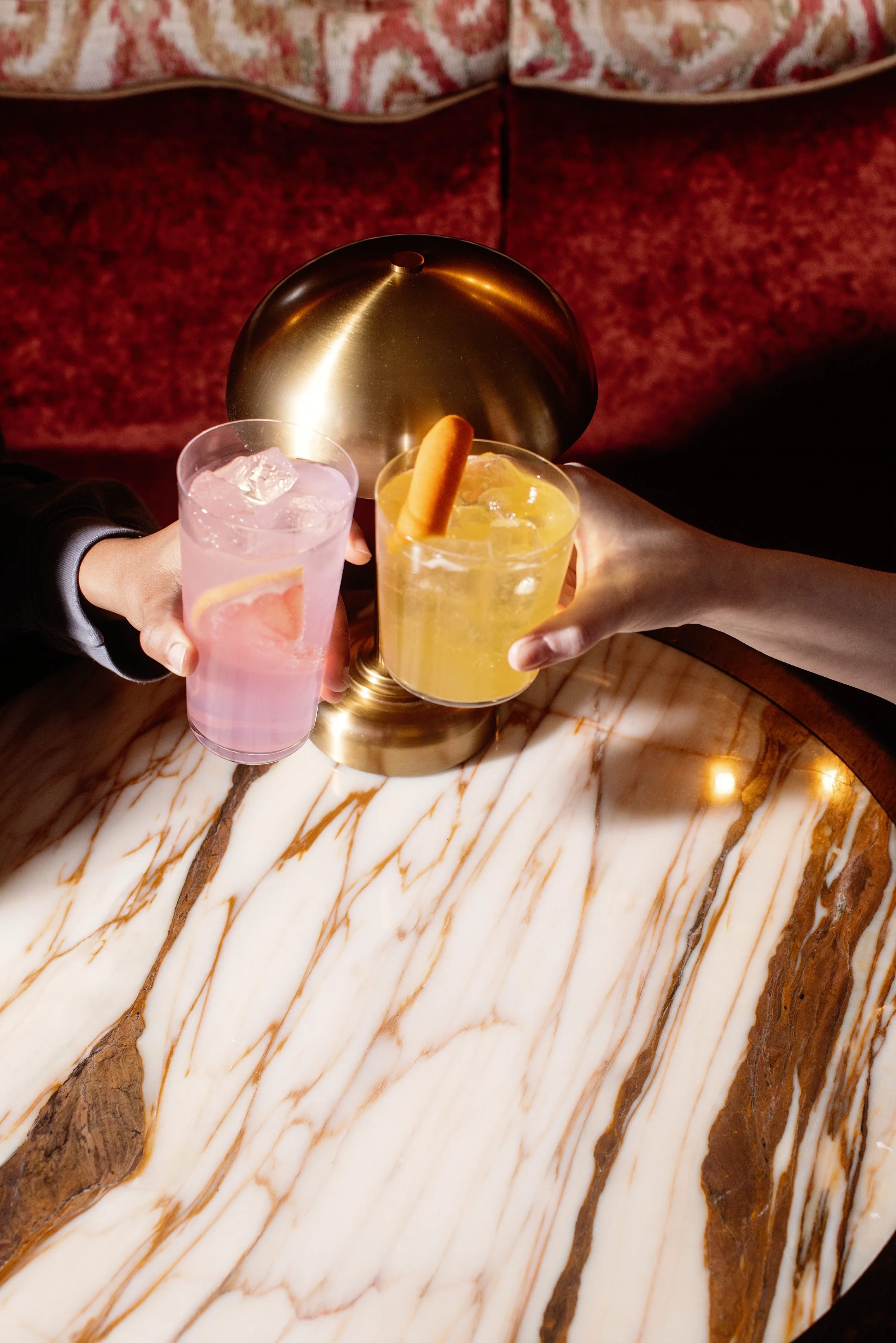 Two people clinking glasses of colorful cocktails, one pink and one yellow, on a marble table with a decorative gold lamp in the background.