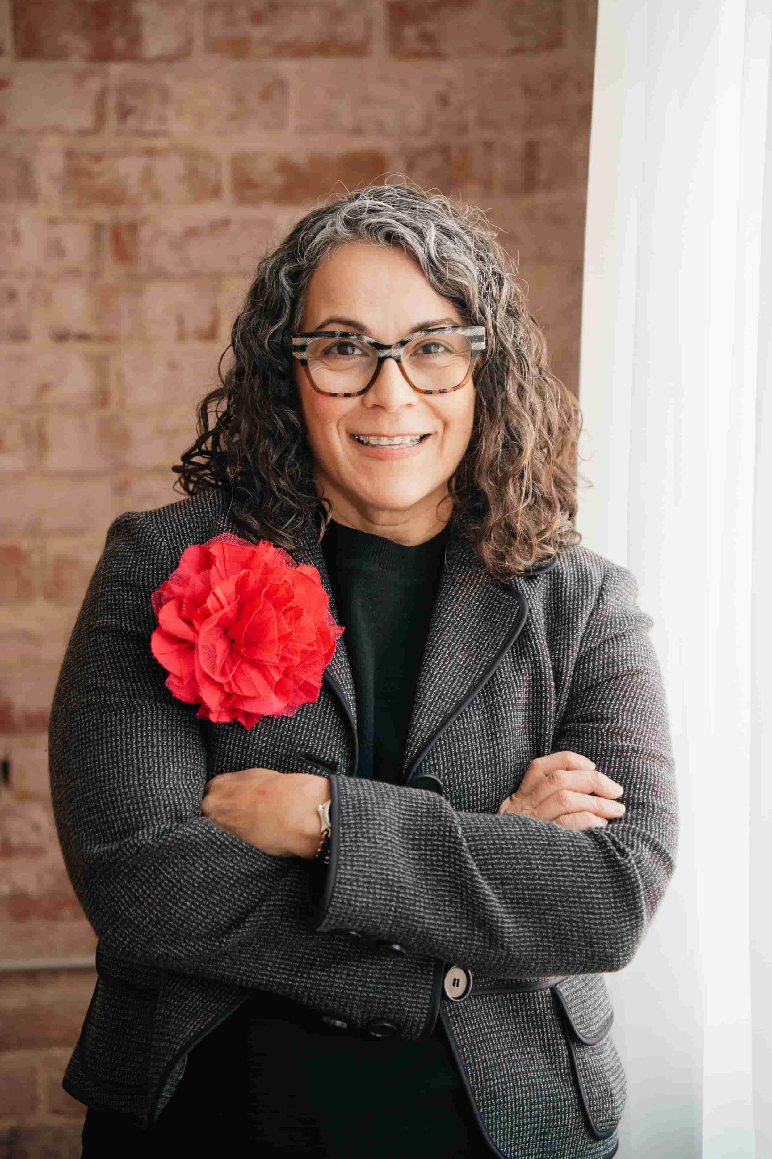 Smiling woman with curly hair and glasses, wearing a blazer with a large red flower, stands with arms crossed by a window.