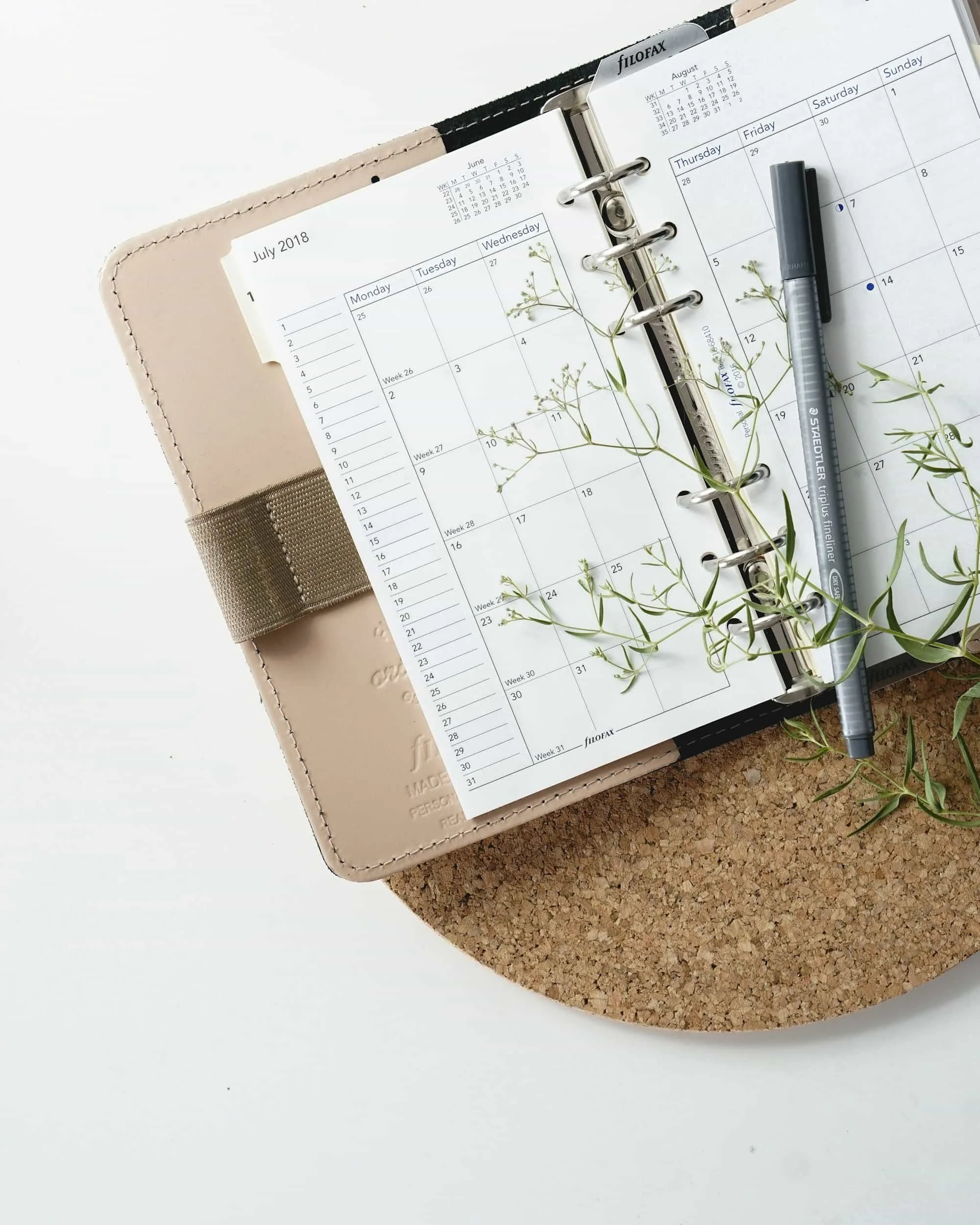 Open planner showing a July 2018 calendar, with a pen and small sprigs of greenery, on a cork mat.
