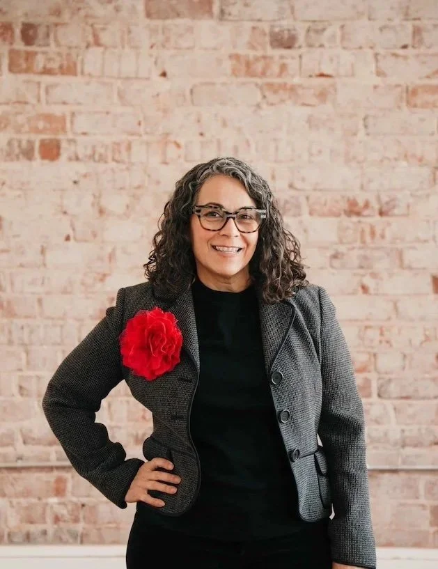 Smiling woman with curly hair and glasses, wearing a blazer with a large red flower, stands with one arm on her hip.