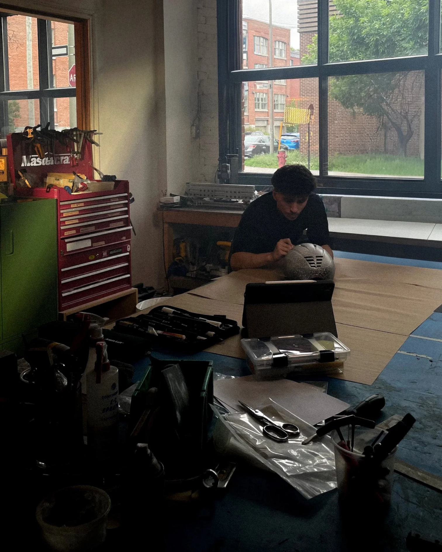 Une personne travaille avec un casque de protection dans une pièce lumineuse, entourée d'outils et de matériel de bricolage sur une grande table, avec fenêtres laissant entrer la lumière naturelle.
