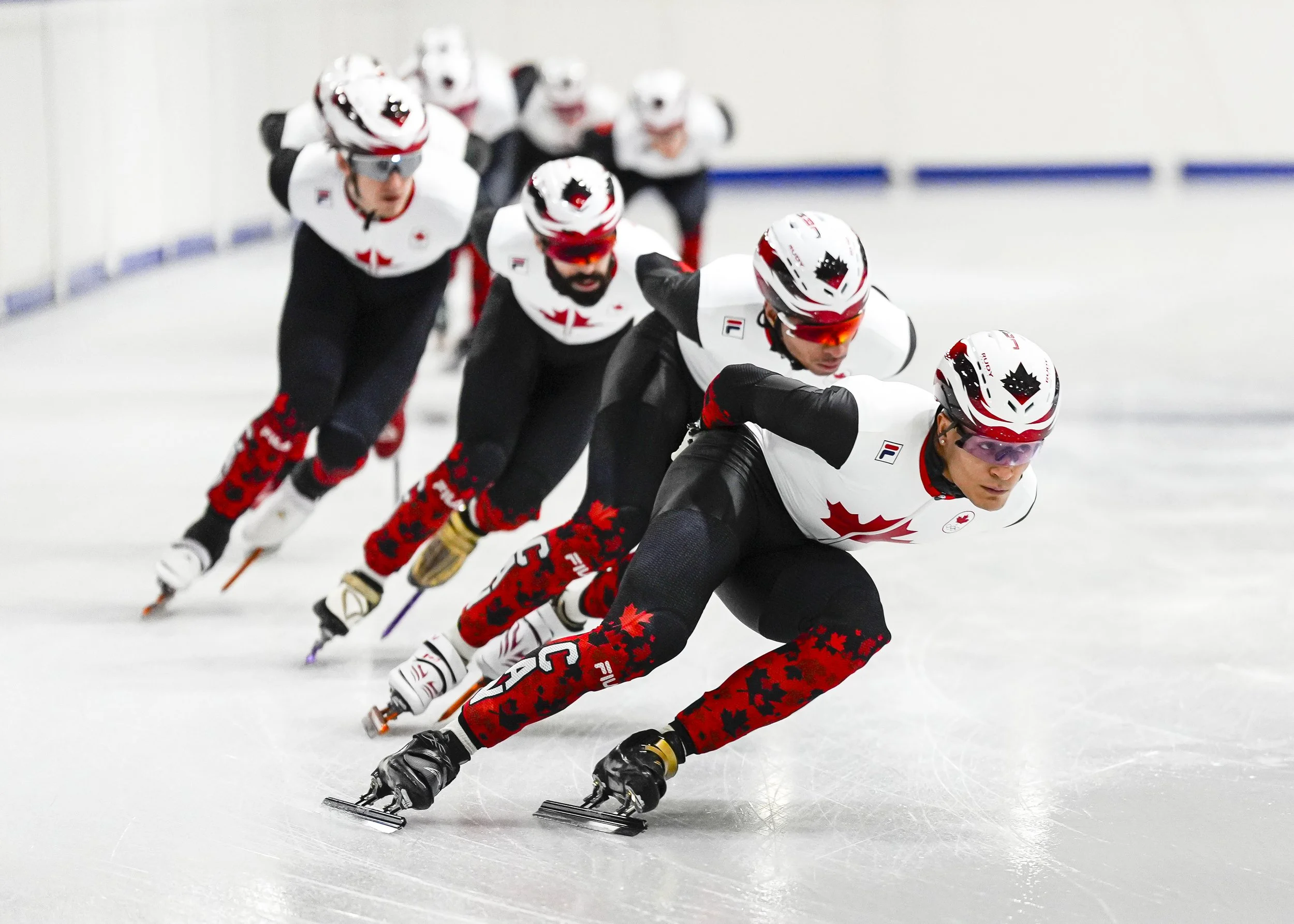 Ce sont des patineurs en ligne en compétition, portant des tenues noires et blanches avec des motifs rouges, des casques blancs avec des visières colorées, et glissant rapidement sur la glace dans une course.