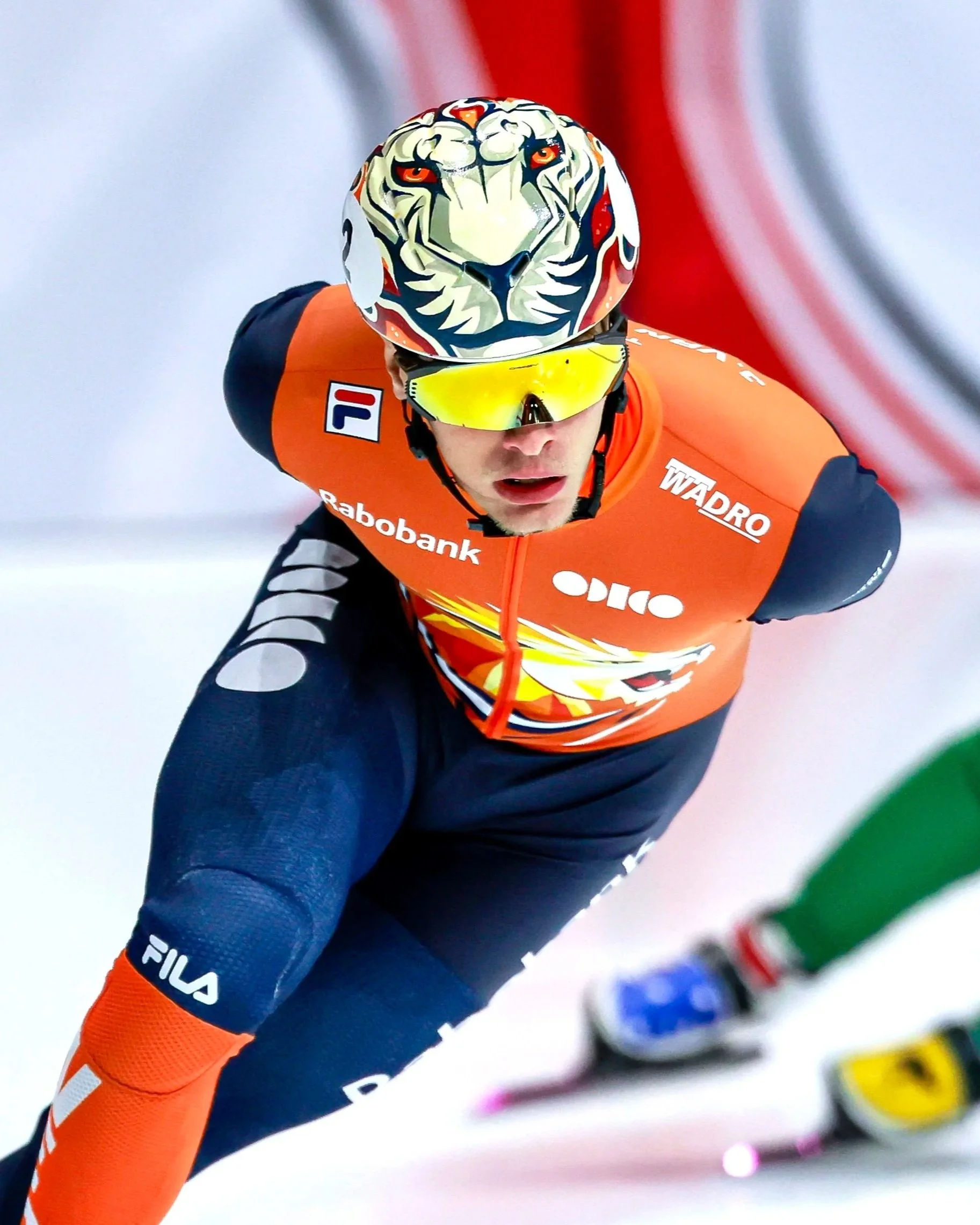Un athlète en course, portant un casque avec un motif de tigre et des lunettes de soleil jaunes, en action sur une piste enneigée, portant un maillot orange avec des logos de sponsors.