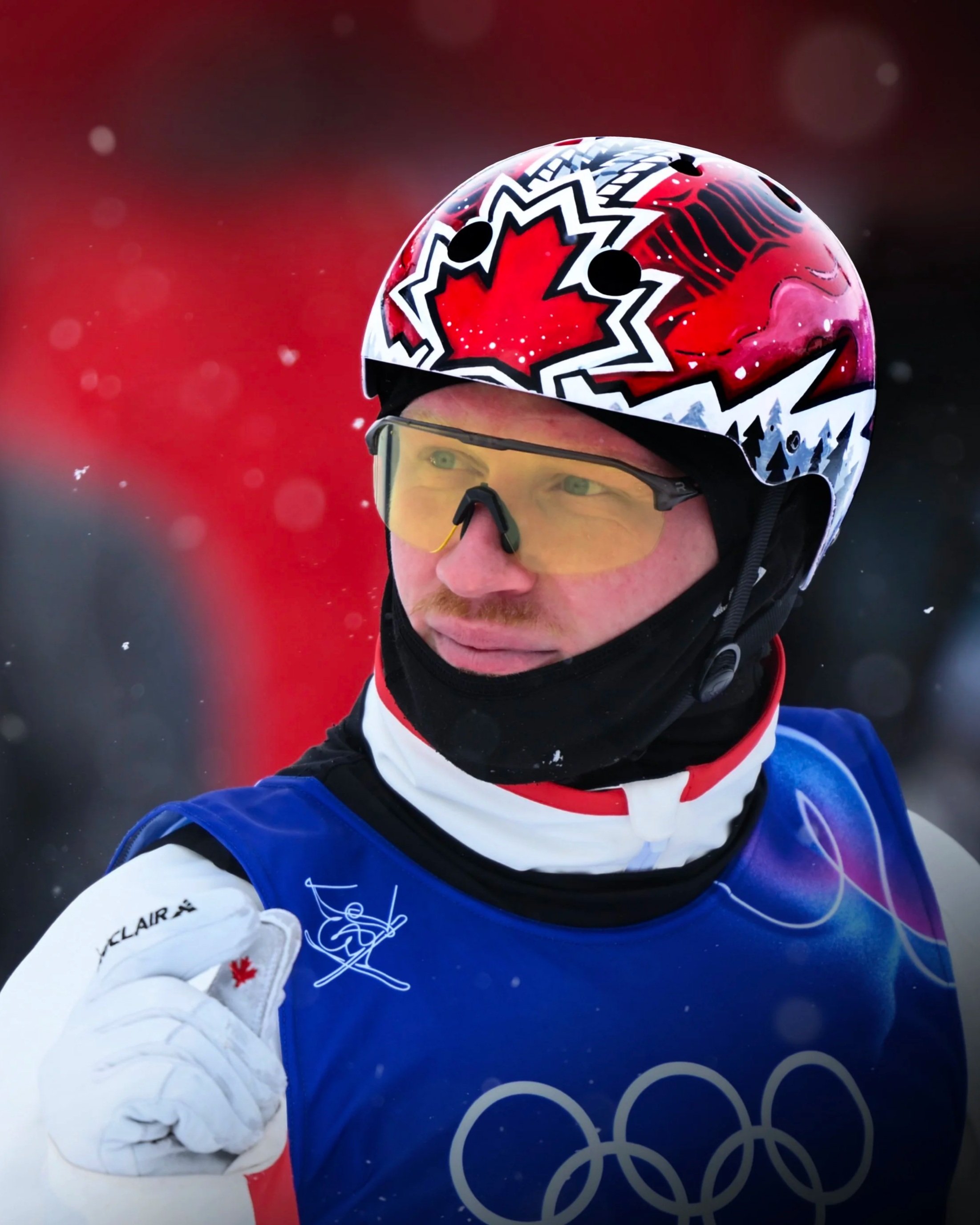 Un athlète de ski alpin portant un casque avec un motif de feuille d'érable, une veste bleue avec le logo des Jeux Olympiques, des lunettes de ski jaunes, et un gant blanc avec une feuille d'érable rouge.