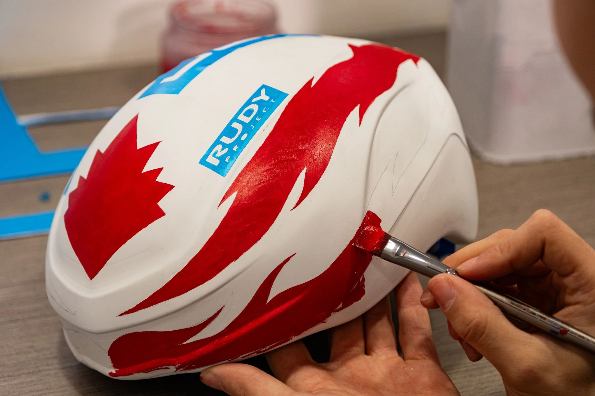 Une personne peint à la main un design rouge et blanc sur un casque de moto blanc, avec une utilisation d'une petite brosse. Le casque comporte aussi un logo bleu avec le mot 'RUDEY'.