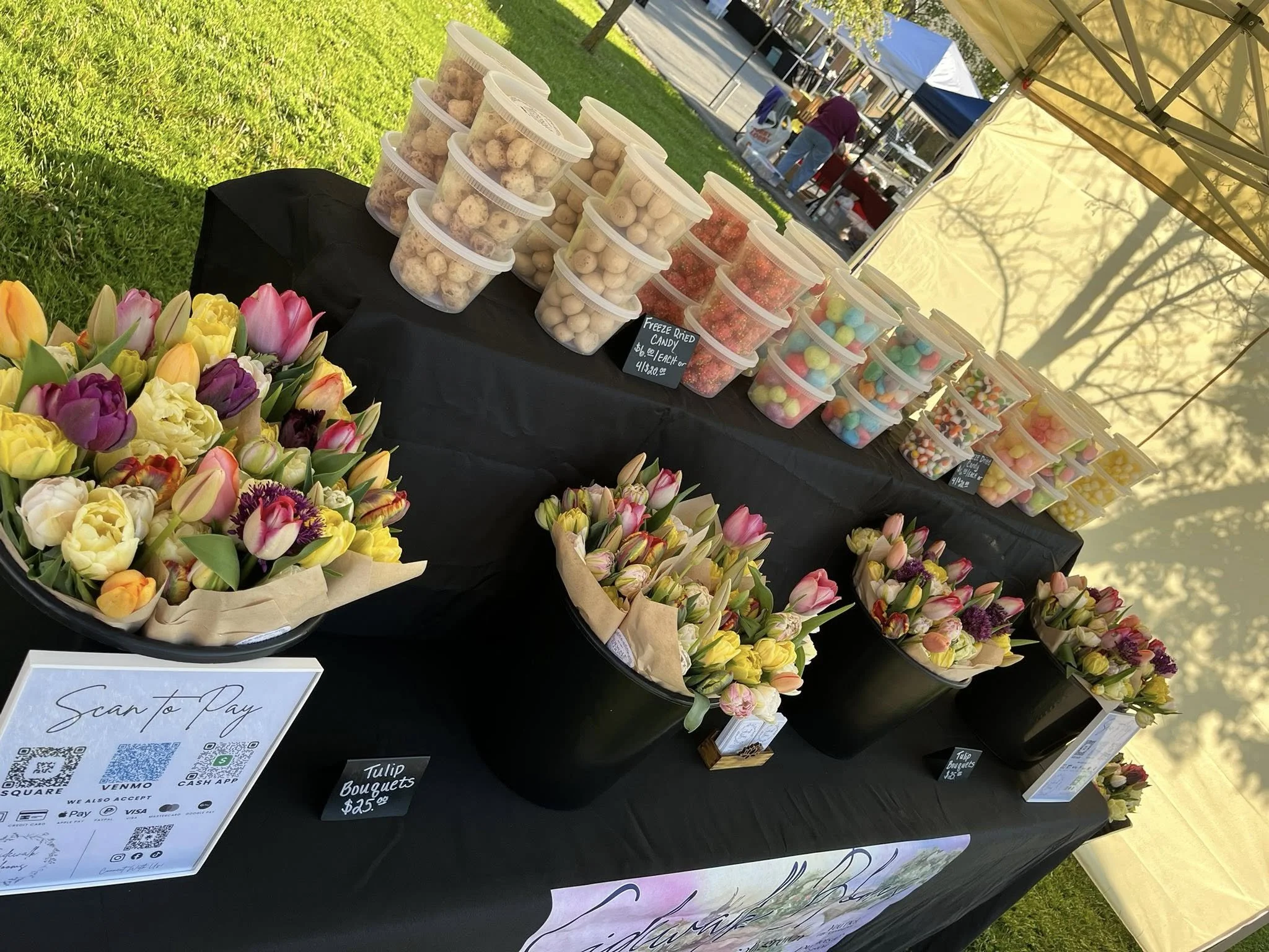 A display table at an outdoor event with colorful tulip bouquets and various containers of candy and sweets, including gummy candies and jellybeans, under a yellow canopy.