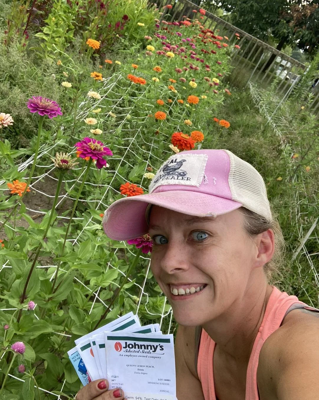 A woman wearing a pink and beige cap and pink tank top is taking a selfie in a flower garden with vibrant orange, yellow, pink, and purple flowers. She holds seed packets from Johnny's Selected Seeds in her hand, smiling at the camera.