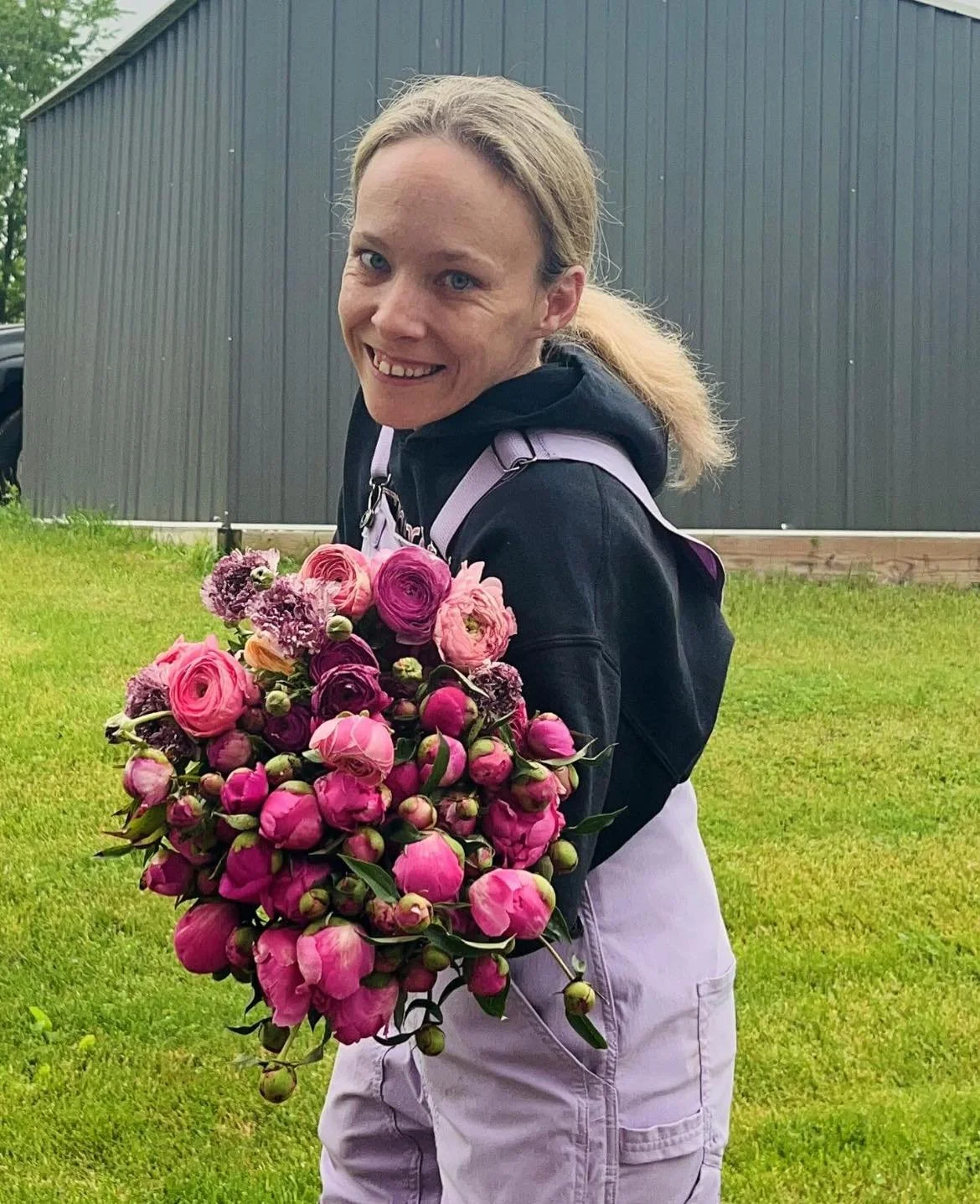 A woman holding a large bouquet of pink and purple flowers, outdoors on a grassy lawn with a dark green building in the background.