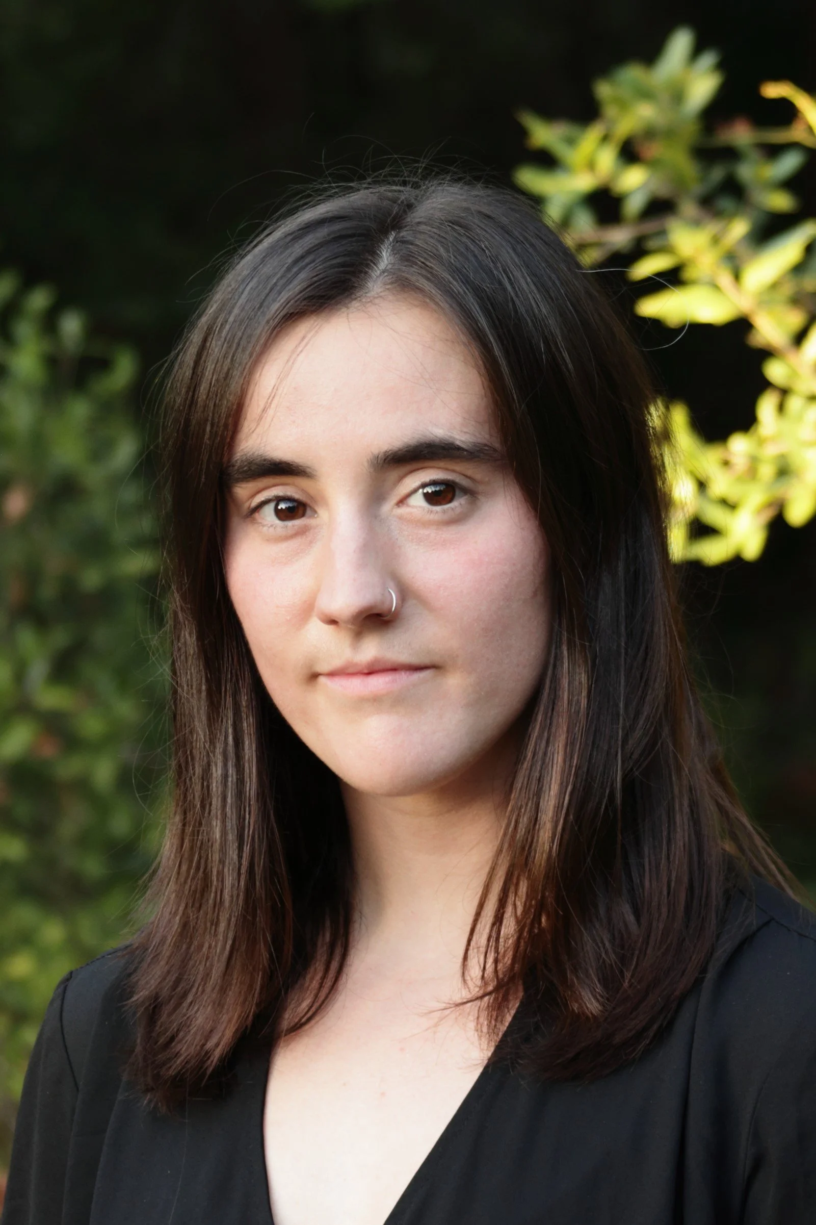 Close-up of a young woman with long, dark brown hair, wearing a silver nose ring and a black top, standing outdoors with greenery in the background.
