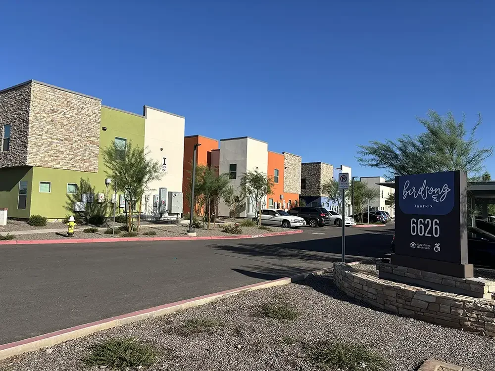 Colorful modern apartment buildings with trees and parked cars in front, signs indicating a residential complex called Birdsong in Phoenix at 6626, under a clear blue sky.