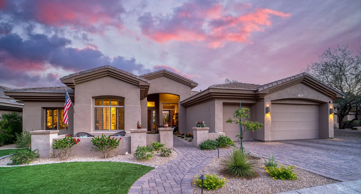 A modern single-story house with a landscaped front yard, a curved brick walkway, an American flag, and illuminated exterior lights at dusk.