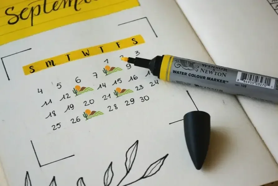 A calendar page for September shows the days of the week and dates, with mid-month marked with small cactus and sunset illustrations. A gray water color marker is placed on the page.