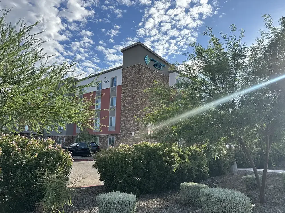 WoodSpring Suites hotel building with a stone and white exterior, green trees and bushes in the foreground, and a partly cloudy sky with sunlight
