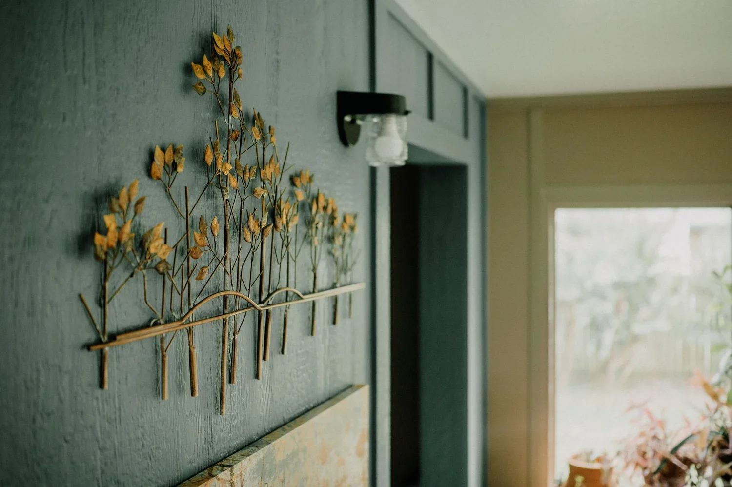 Decorative metal wall art of a tree with yellow leaves in a room with a wall-mounted light fixture and a large window with potted plants outside.