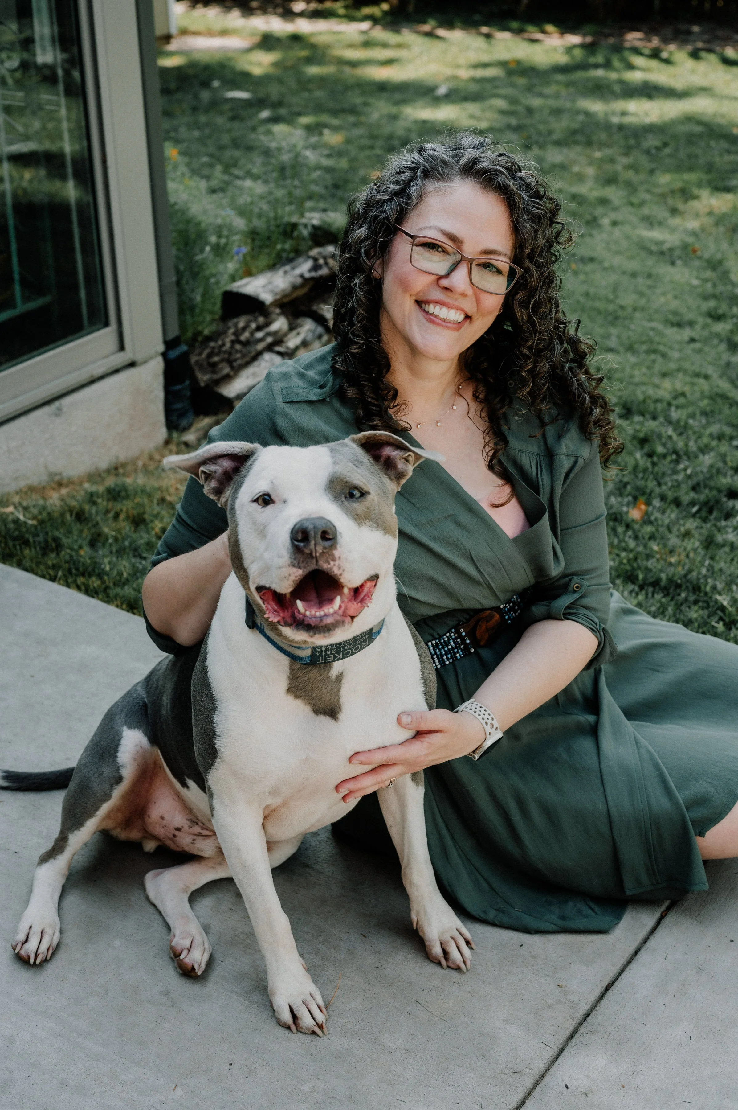 A woman with curly dark hair, glasses, and a green dress sitting outdoors on a concrete patio, smiling, with a happy gray and white pitbull sitting beside her, also smiling.
