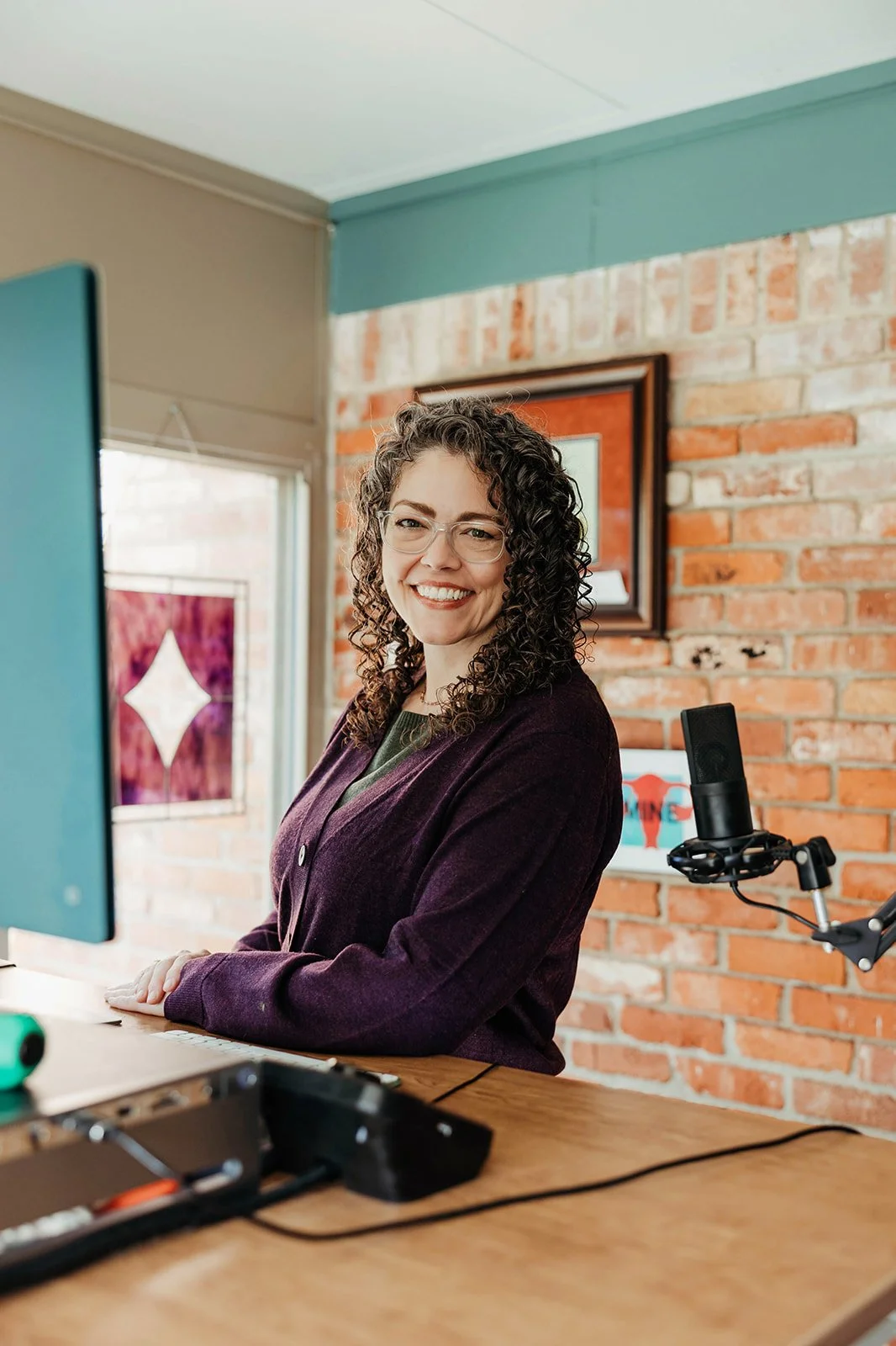 A woman with curly hair and glasses sitting at a desk with a microphone and computer, smiling in a room with a brick wall and artwork.