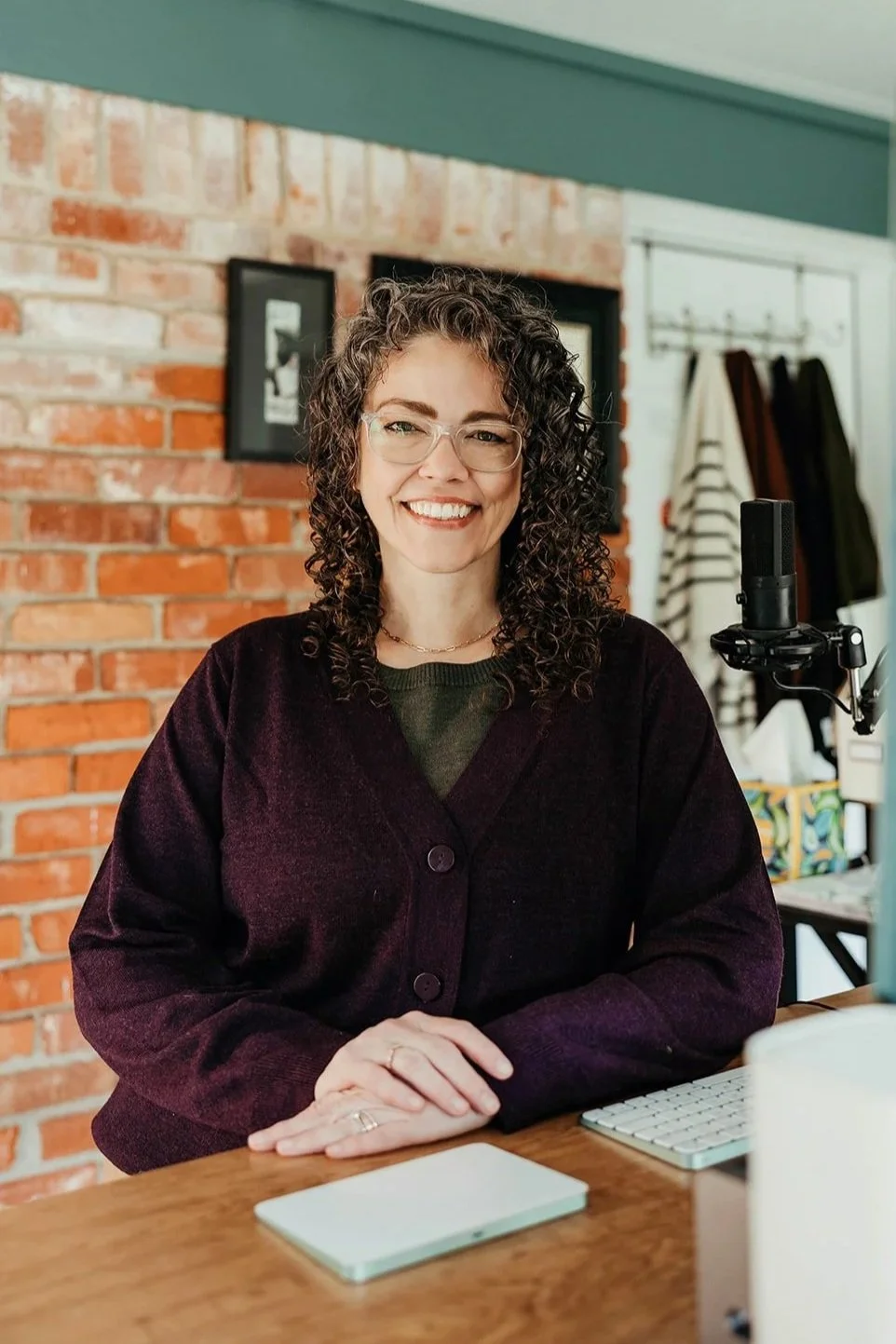 A woman with curly hair, glasses, and a dark sweater sitting at a desk with a microphone, a keyboard, and a tablet in front of her, smiling in a room with a brick wall and framed photos in the background.