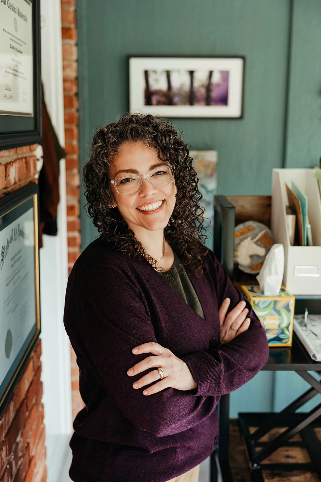 A woman with curly hair and glasses smiling, standing in an office or professional setting with framed certificates and artwork in the background.