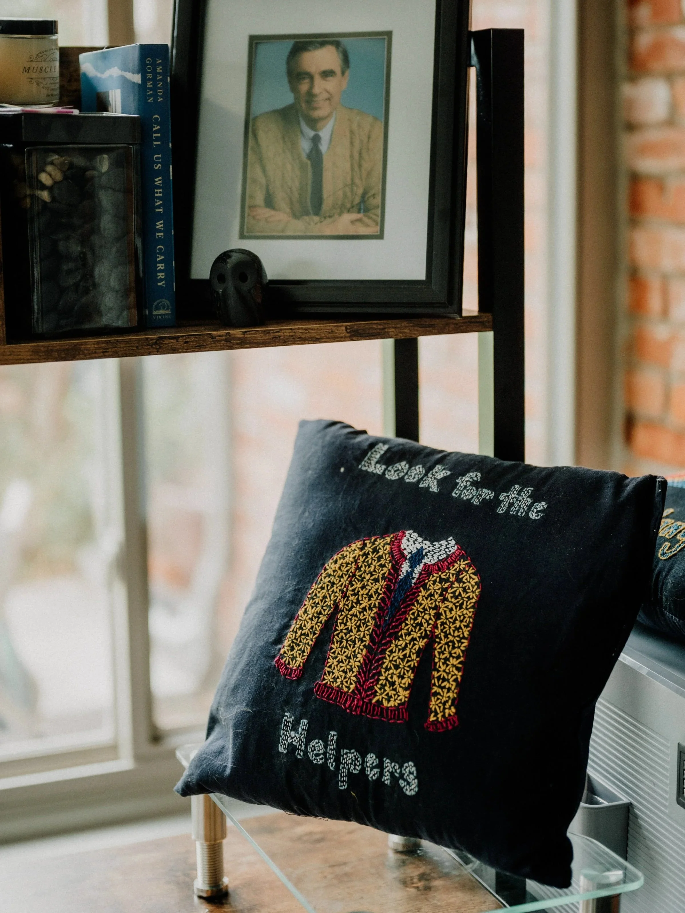 A black pillow with an embroidered yellow and red jacket and the words "Look for the Helpers" on it, resting on a surface in a room with a view of a window and a brick wall outside.