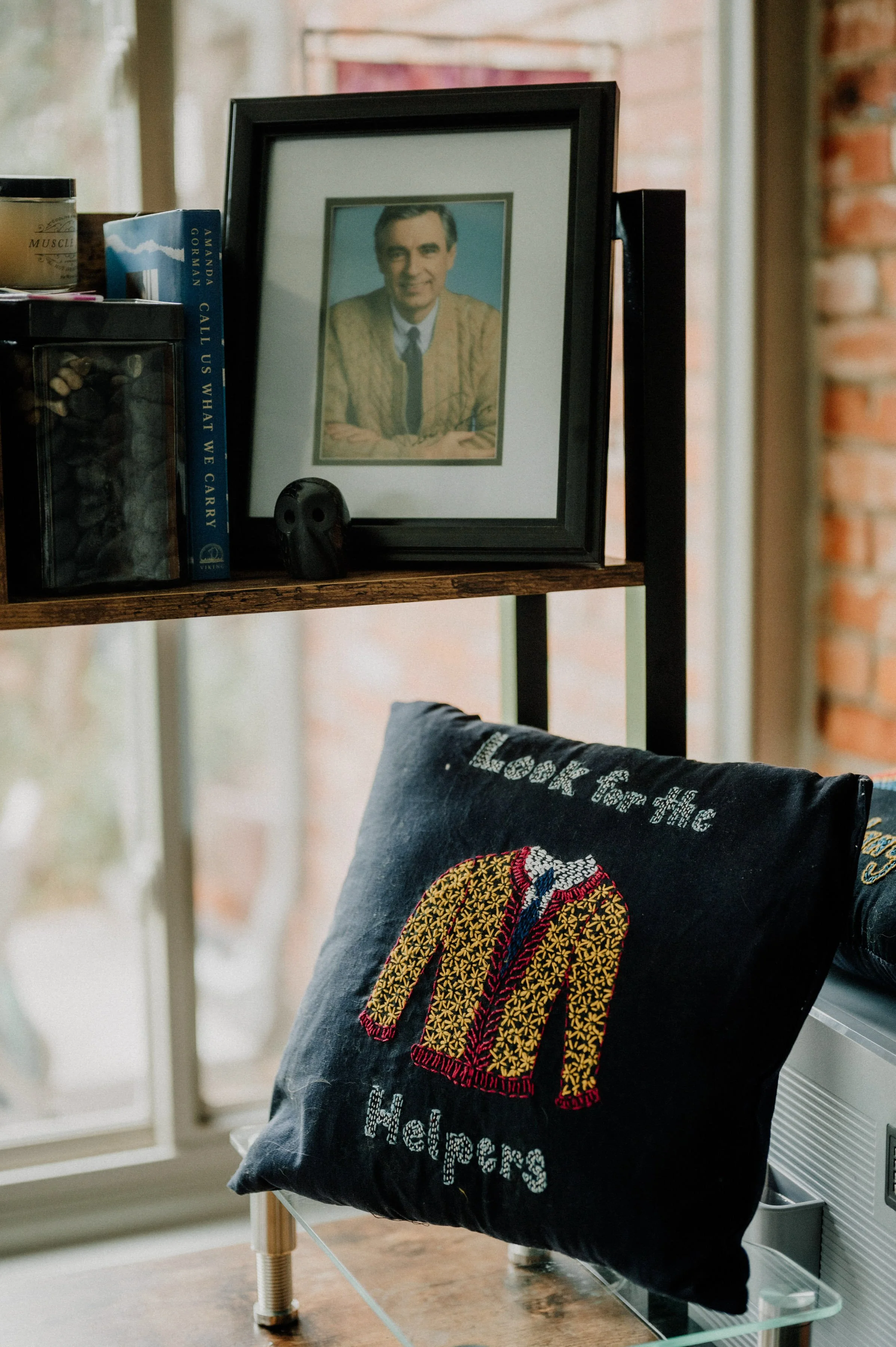 An autographed photo of Mr. Rogers, placed on a wooden shelf with books and a small container. In front of the shelf, a black pillow with an embroidered yellow and red jacket and the words "Look for the Helpers"  is on display.