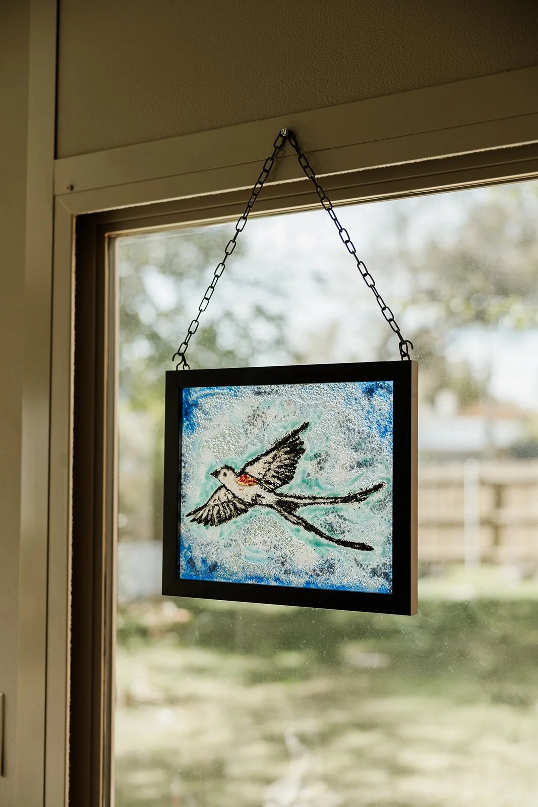 Framed artwork of a bird with outstretched wings, hanging on a chain in front of a window.