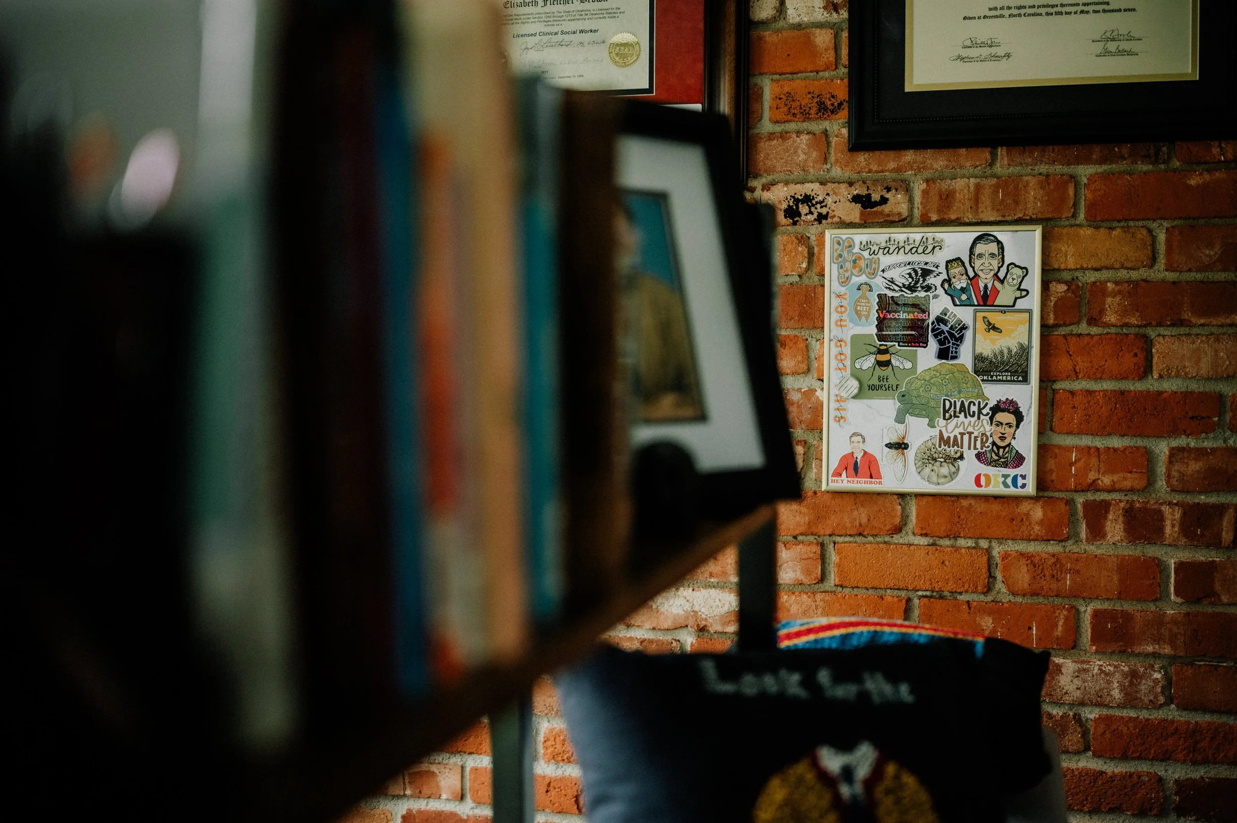 Partially visible bookshelf with books in the foreground, a brick wall with framed documents, and a colorful collage poster on the wall.