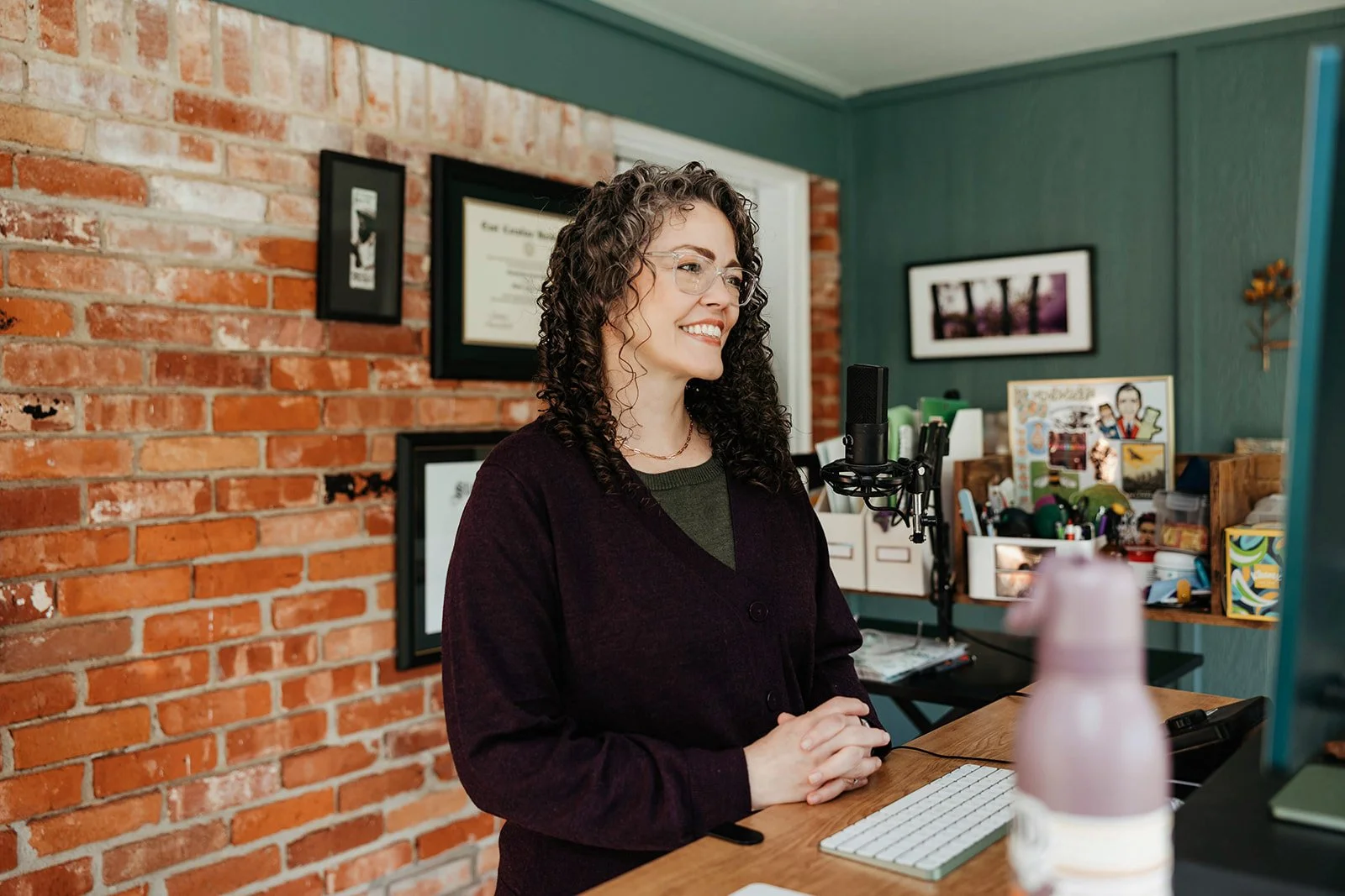 Woman with curly hair and glasses smiling at a desk with a computer keyboard and microphone in a home office with brick wall and framed pictures
