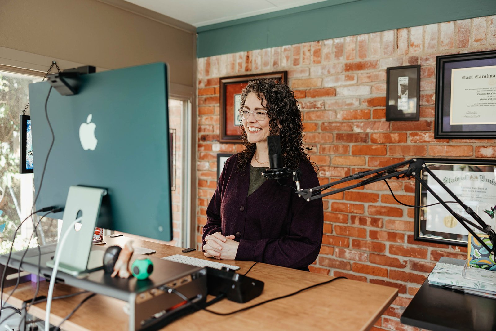 A woman with curly hair and glasses smiling during a video call or recording session, sitting at a desk with a desktop computer, microphone, and various items in a room with brick walls and framed certificates on the wall.