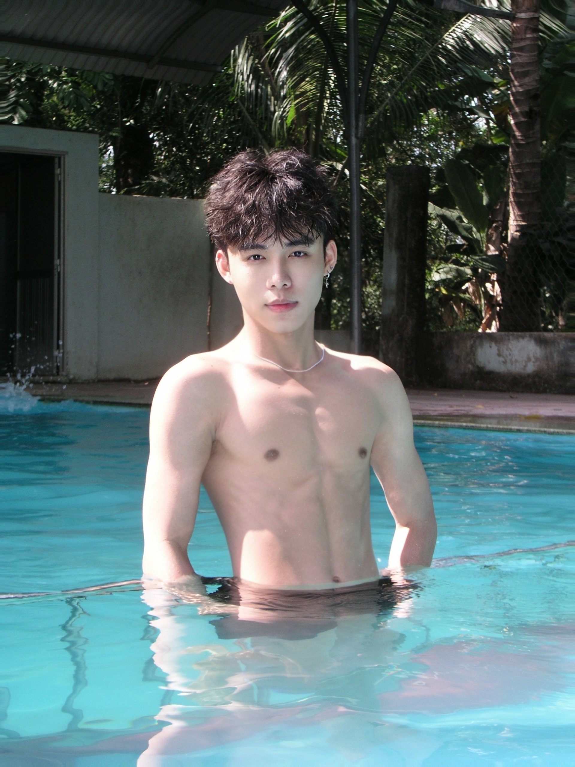 Young man with dark, tousled hair on a swimming pool, wearing earrings and a necklace, with a background of tropical plants and a shaded structure.